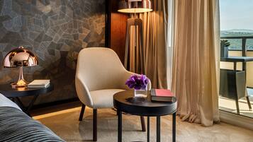 A hotel room with a stone-patterned wall, a round table with a lamp, a chair, and a view of the outdoors through a window with curtains.