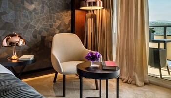 A hotel room with a stone-patterned wall, a round table with a lamp, a chair, and a view of the outdoors through a window with curtains.