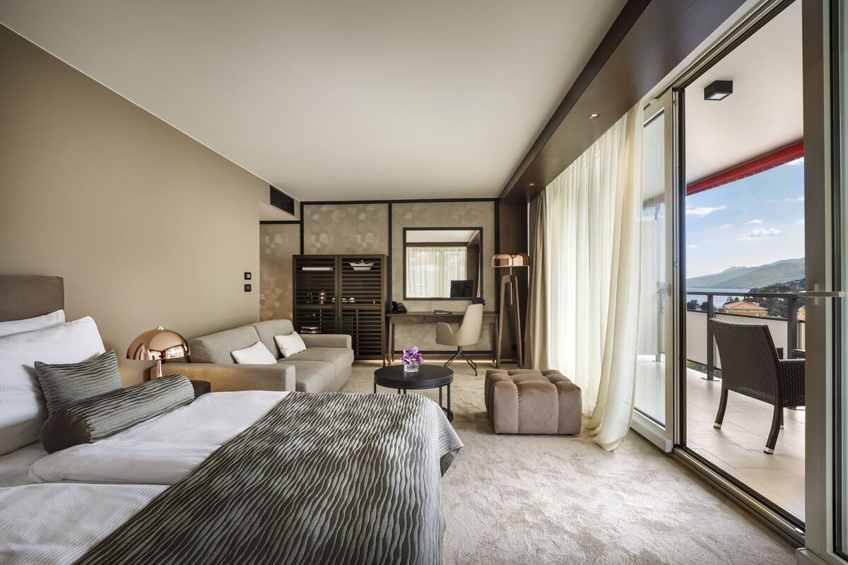 A modern hotel room with a large bed, a sofa, a desk, and a balcony with a view.