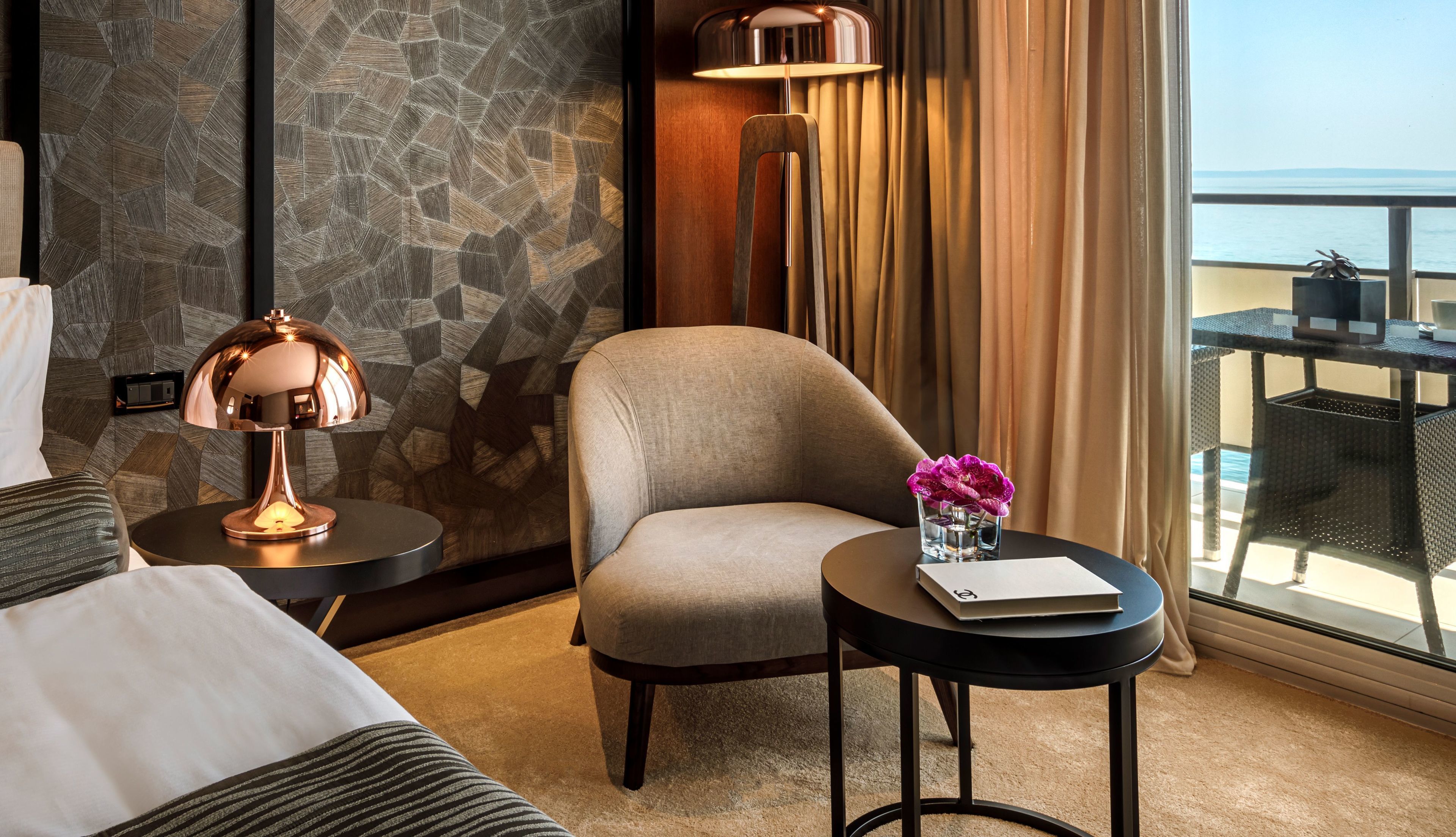 A hotel room with a stone wall, a lamp, a chair, a small table with a vase of flowers, and a view of the sea through large windows.