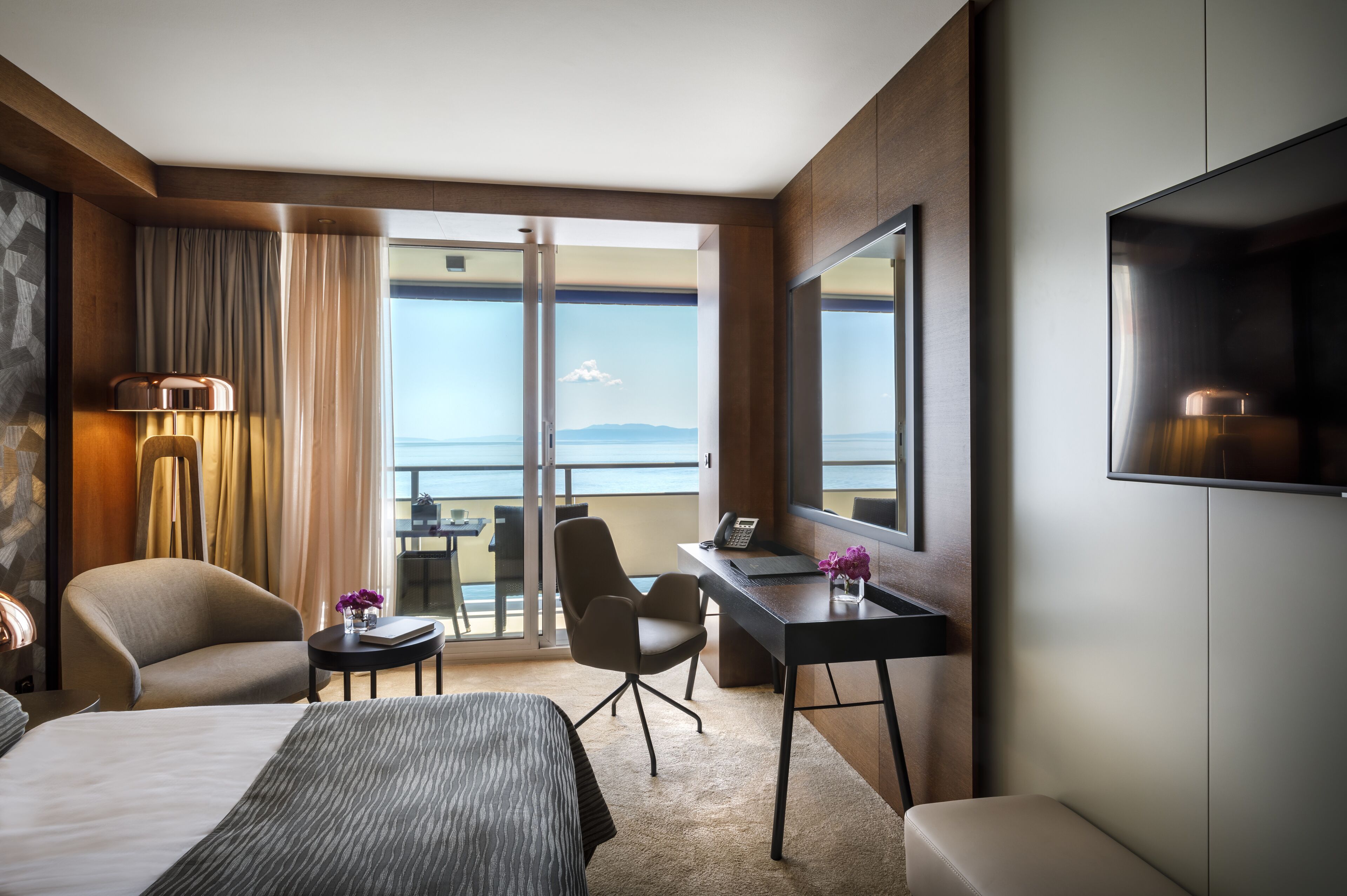 Een moderne hotelkamer met een groot bed, een bureau met een computer, een stoel en uitzicht op zee vanaf een balkon.