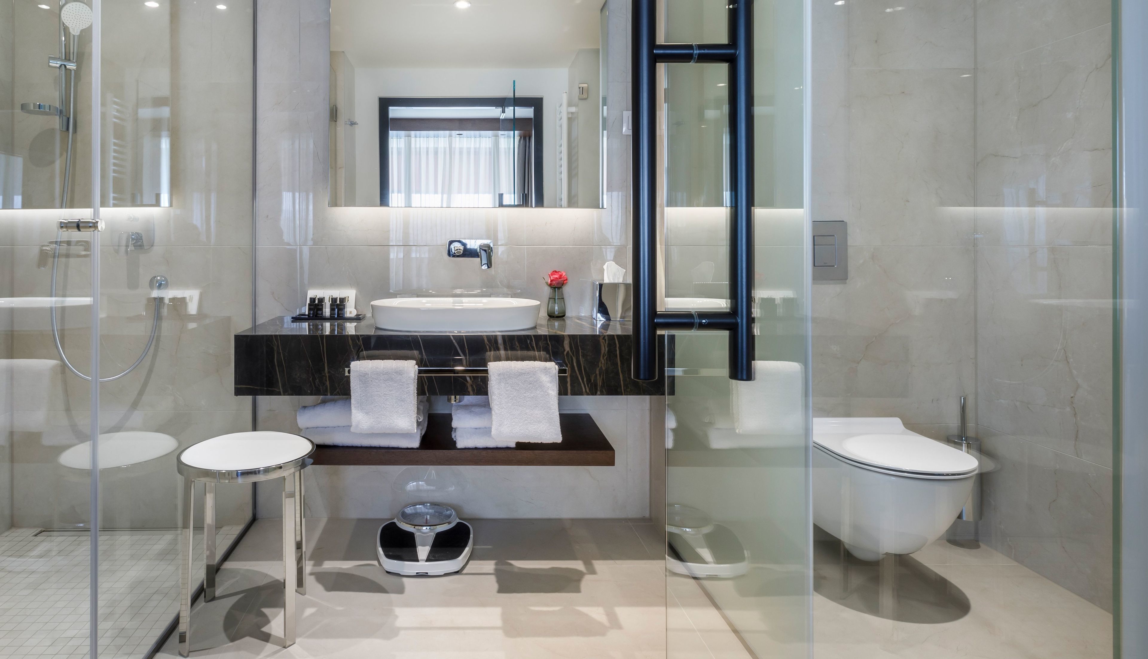 Een moderne badkamer met een douchecabine van glas, een wastafel met een marmeren blad, twee witte toiletten en een weegschaal op de vloer.