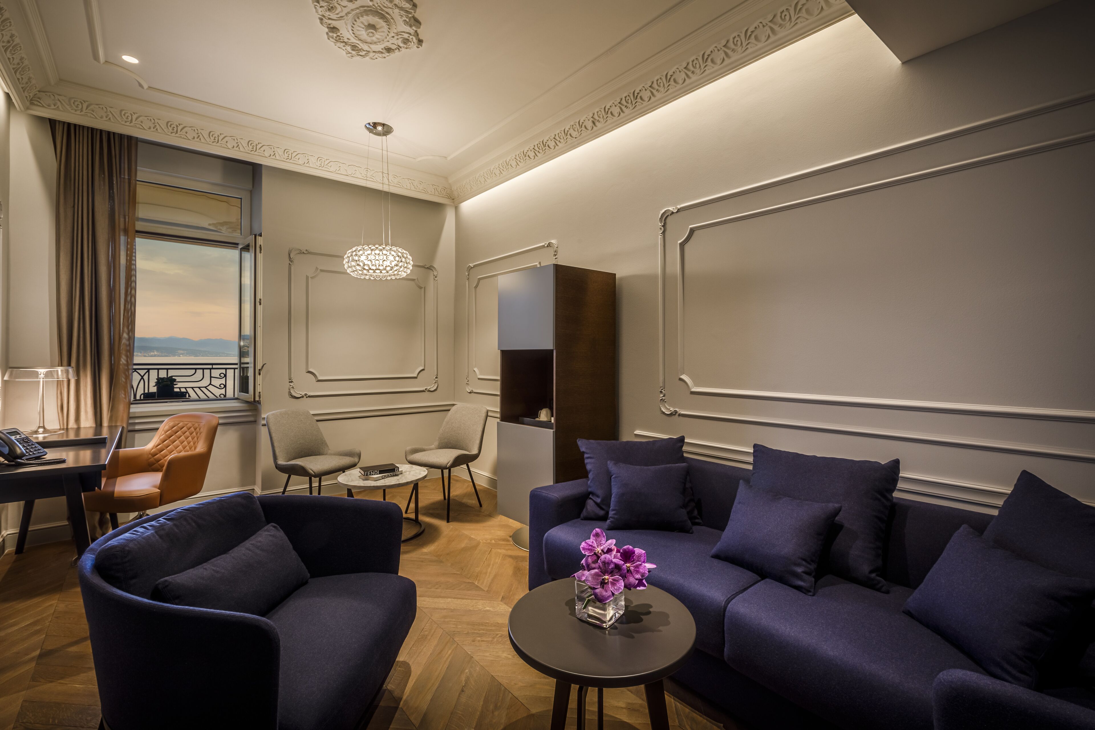 A modern hotel room with a sofa, armchairs, a small table with a vase of flowers, and a view of the cityscape through a window.
