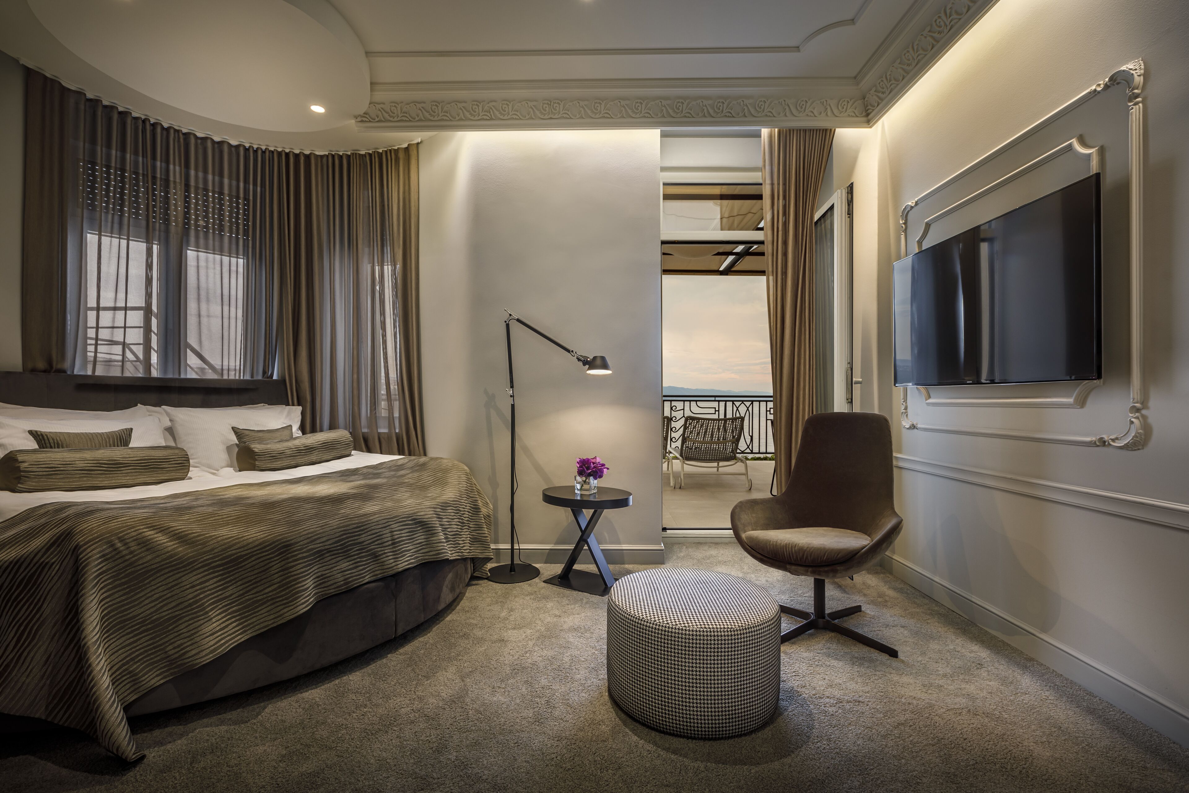 A modern hotel room with a large bed, a round ottoman, a desk with a lamp, a TV, and a view of the outdoors.