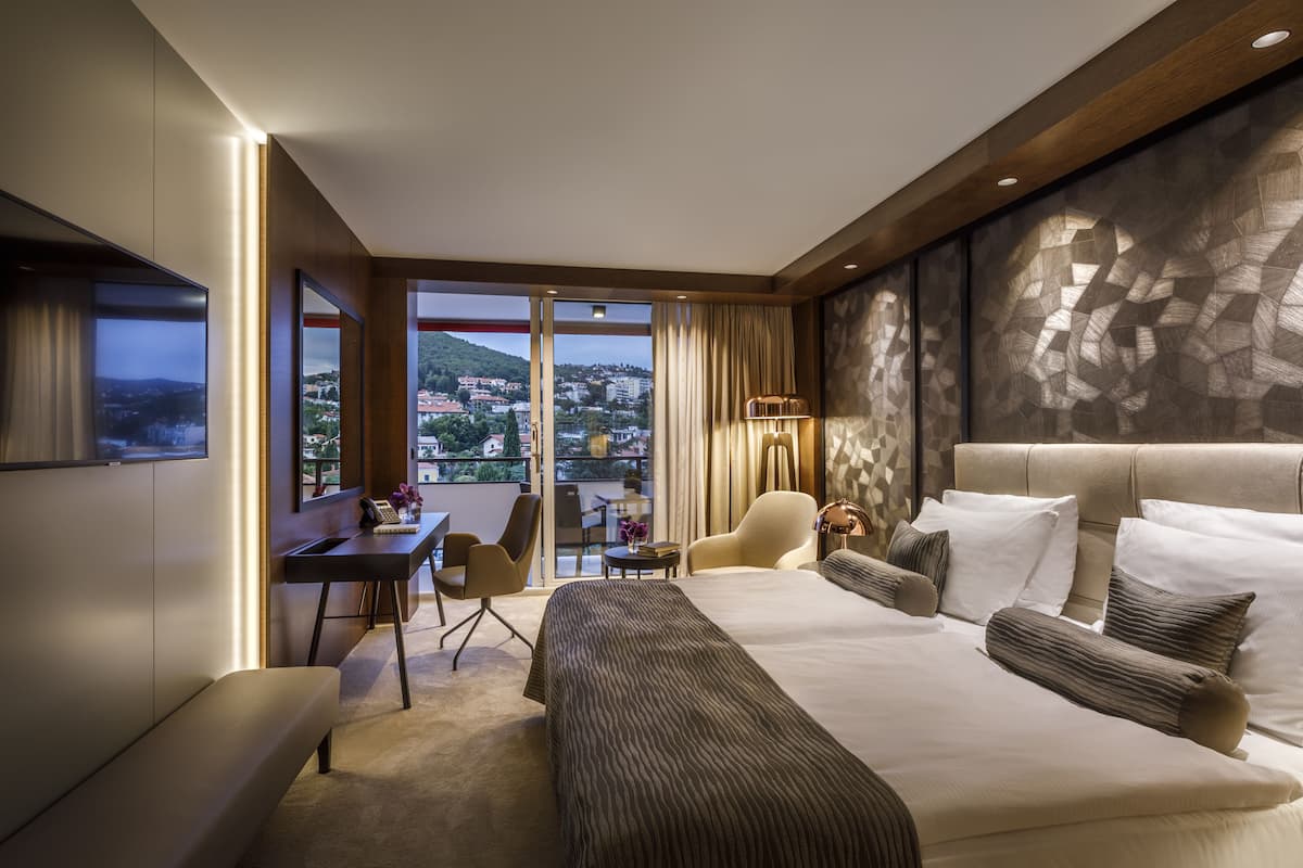 A modern hotel room with a large bed, a desk, and a chair. There is a view of a cityscape through the window.