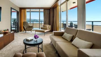 A modern hotel room with a large balcony, a sofa, a chair, and a small table with a book and flowers.
