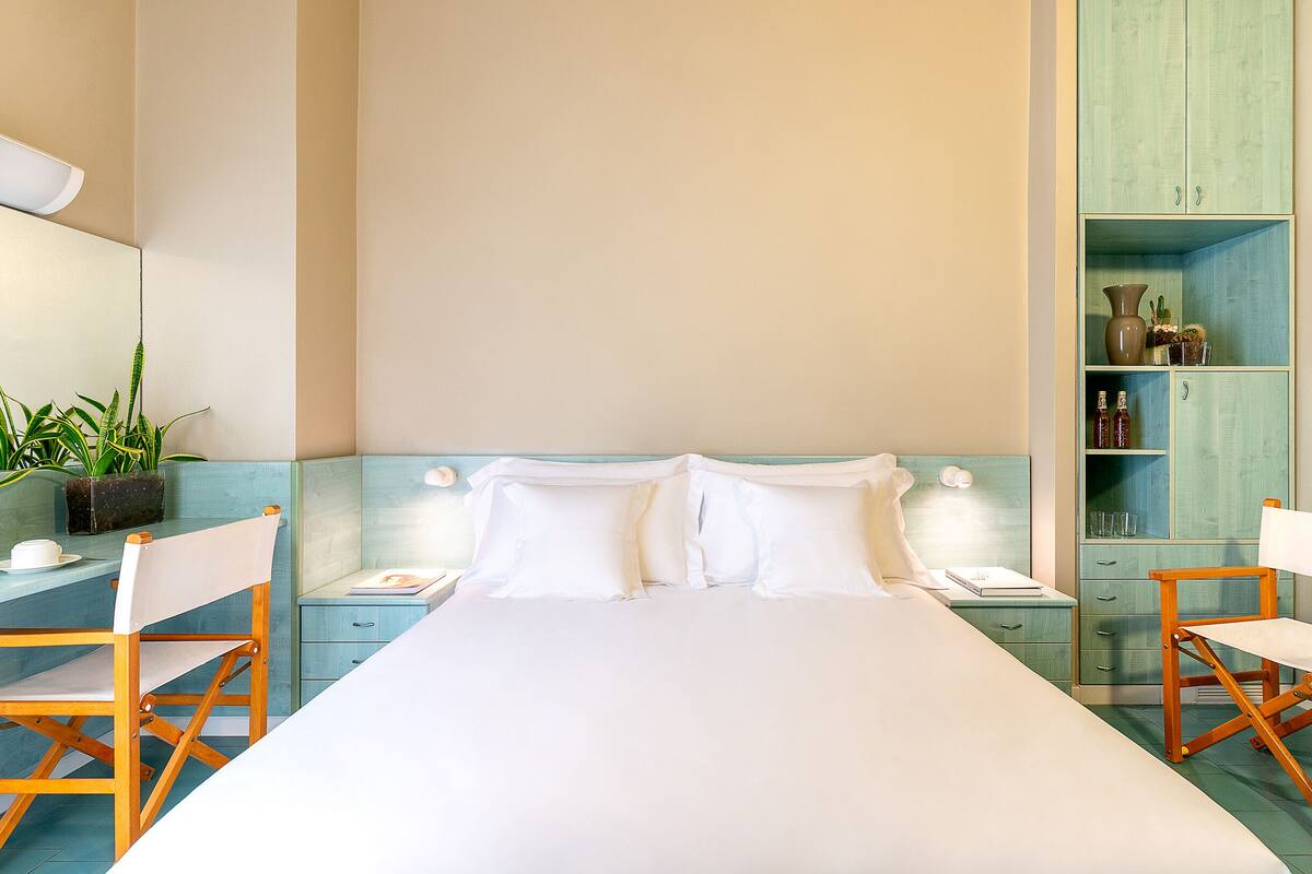 Una camera d'albergo con un letto grande, due sedie di legno, un tavolino con un bollitore, una pianta in vaso e un minibar.