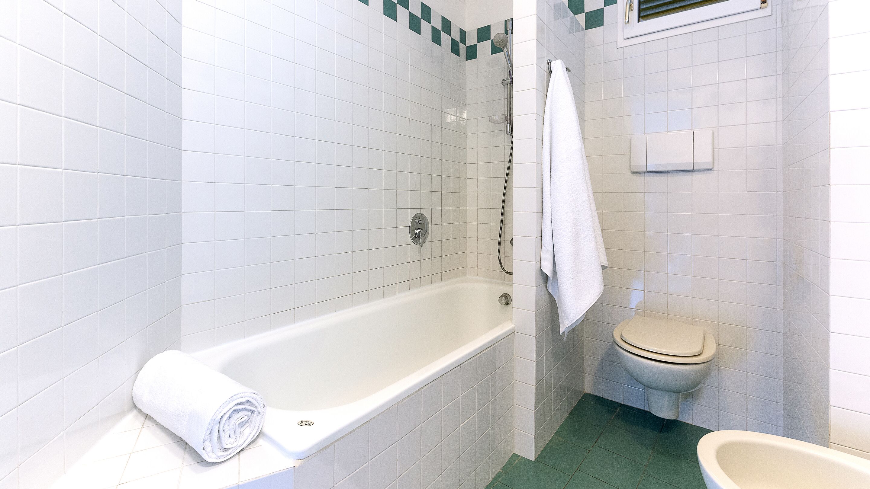 A bathroom with white tiled walls, a white bathtub, a toilet, and a green floor.