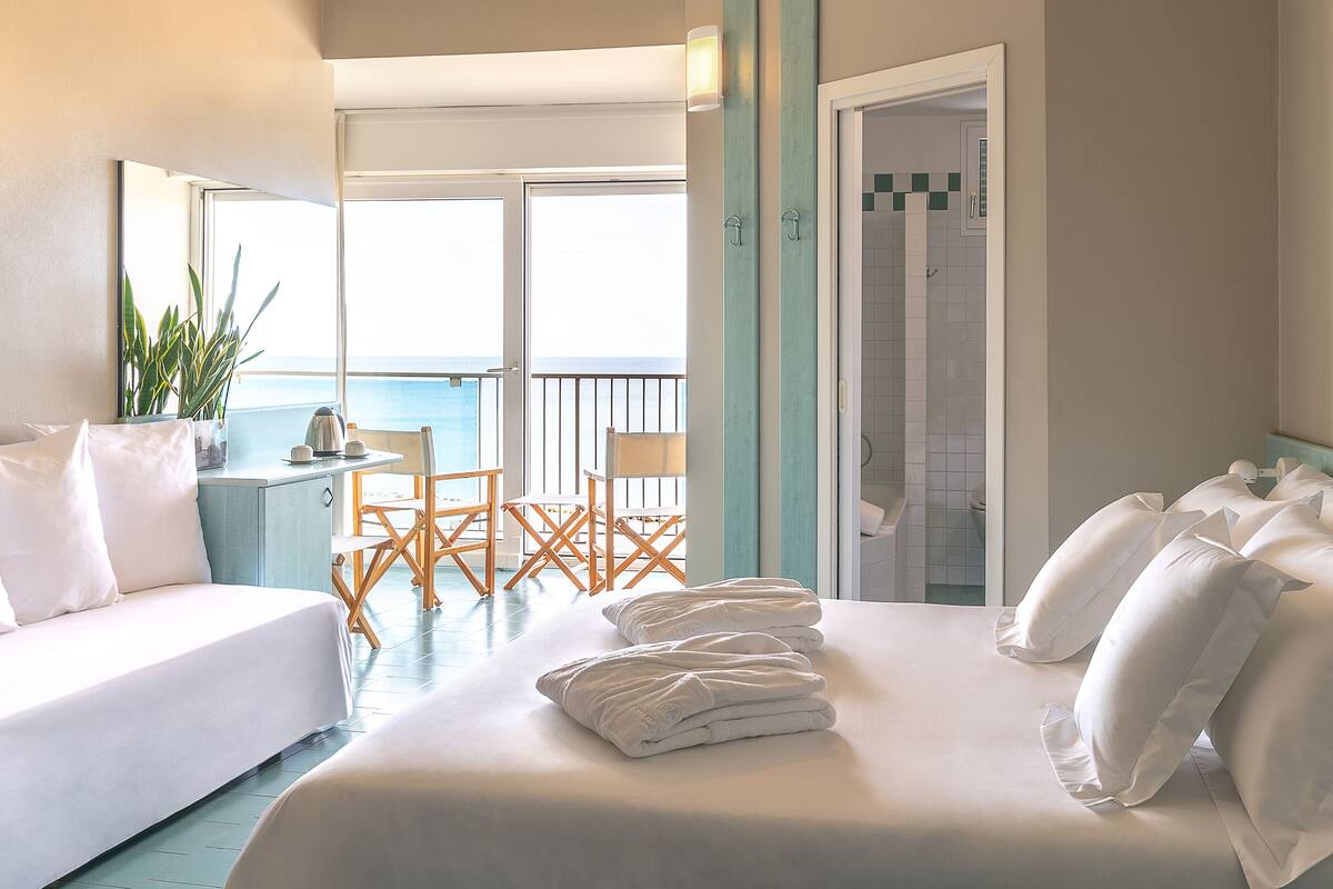 Una camera d'albergo con un letto, un balcone con sedie, un piccolo tavolo con un bollitore e una cabina doccia.