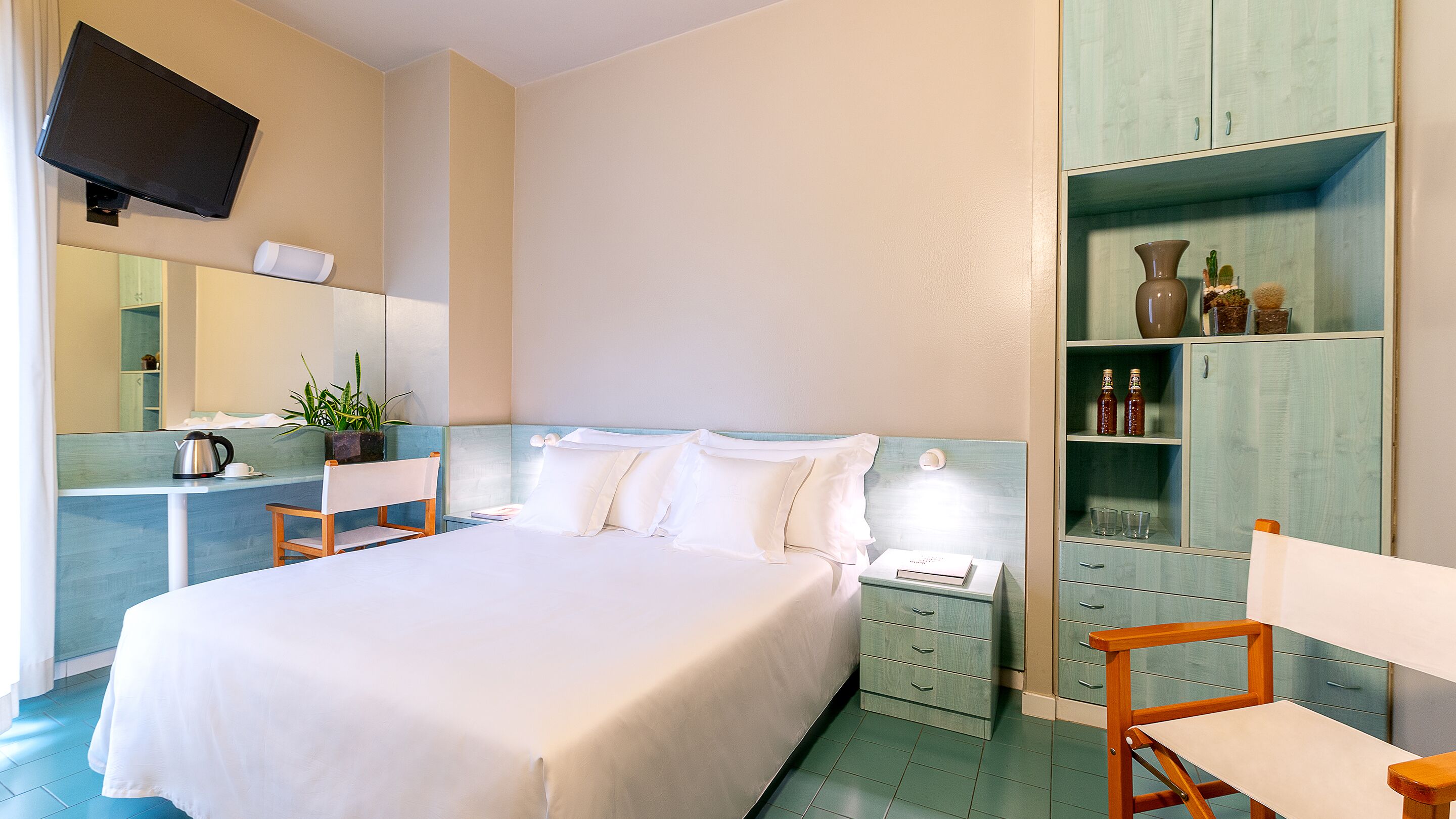 A hotel room with a bed, a bedside table, a nightstand, a mirror, a television, a wardrobe, and a small seating area.