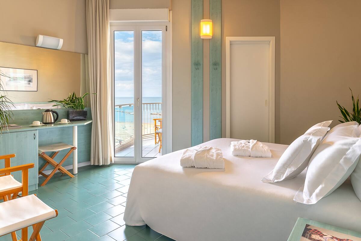 Una camera d'albergo con un letto, una scrivania, una sedia, un balcone vista e una porta.
