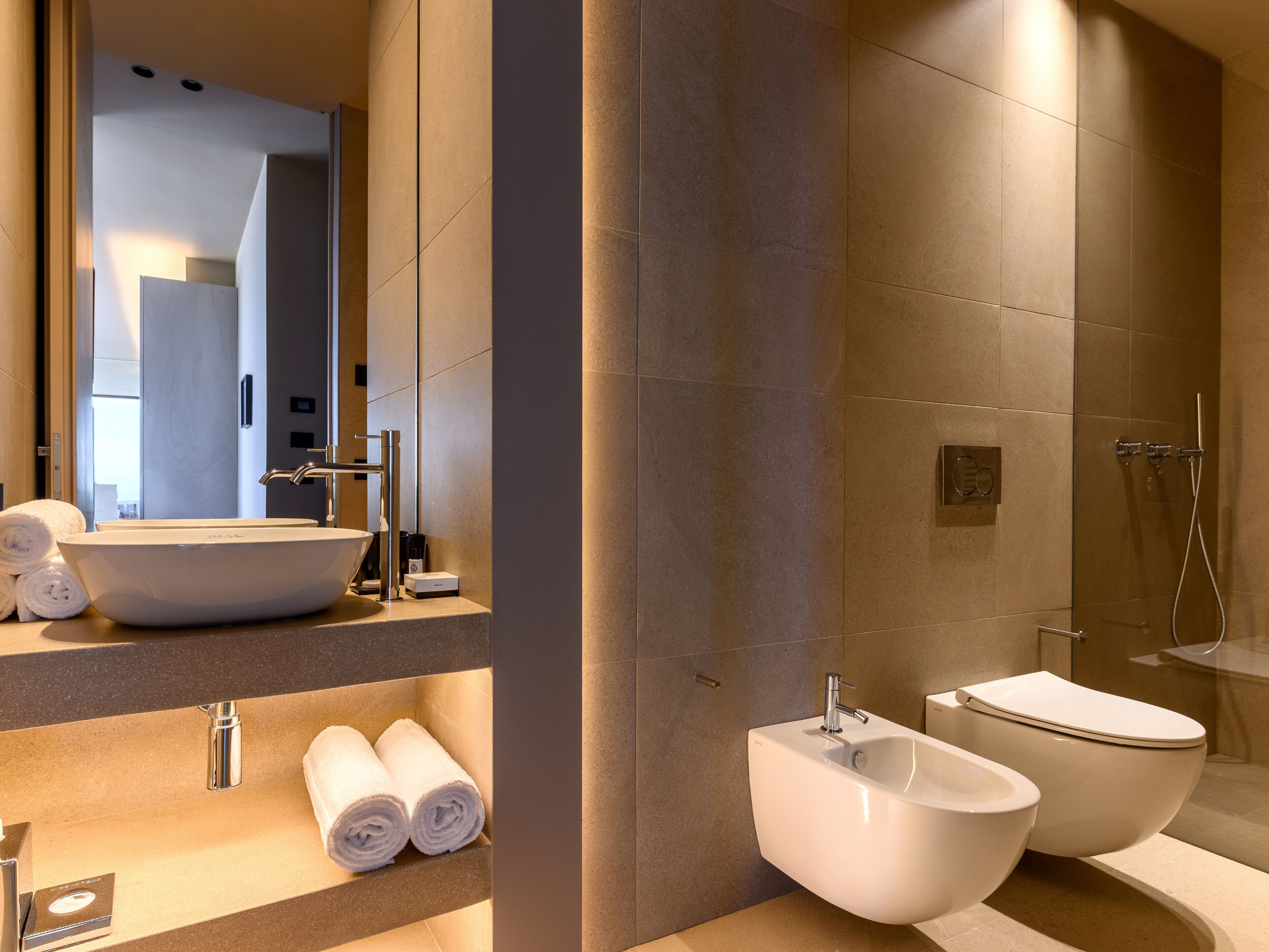 A modern bathroom with a large vessel sink, a bidet, and a toilet. There are towels and a soap dispenser on the counter.