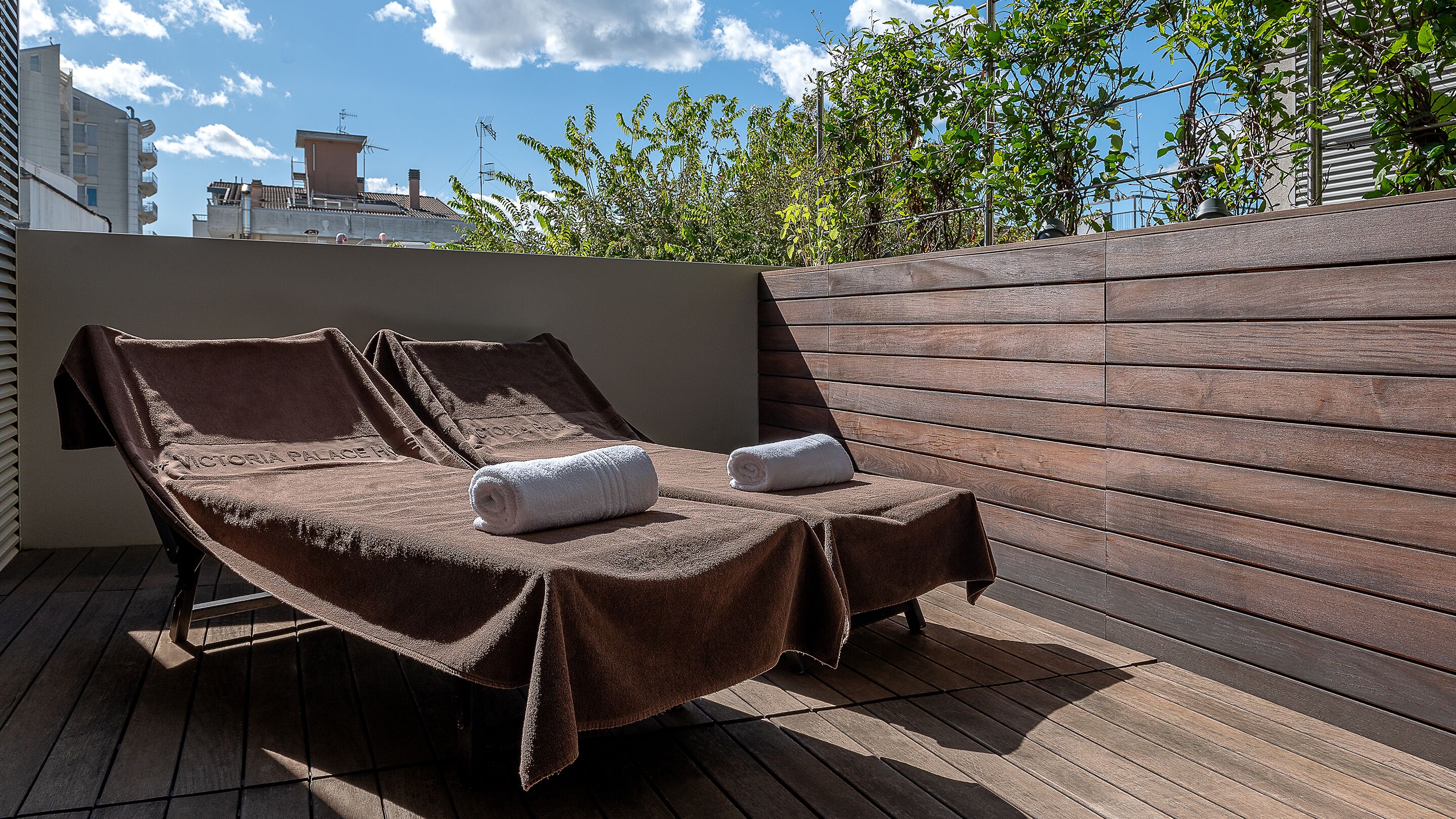 A sun lounger with towels on a wooden deck, overlooking a cityscape.