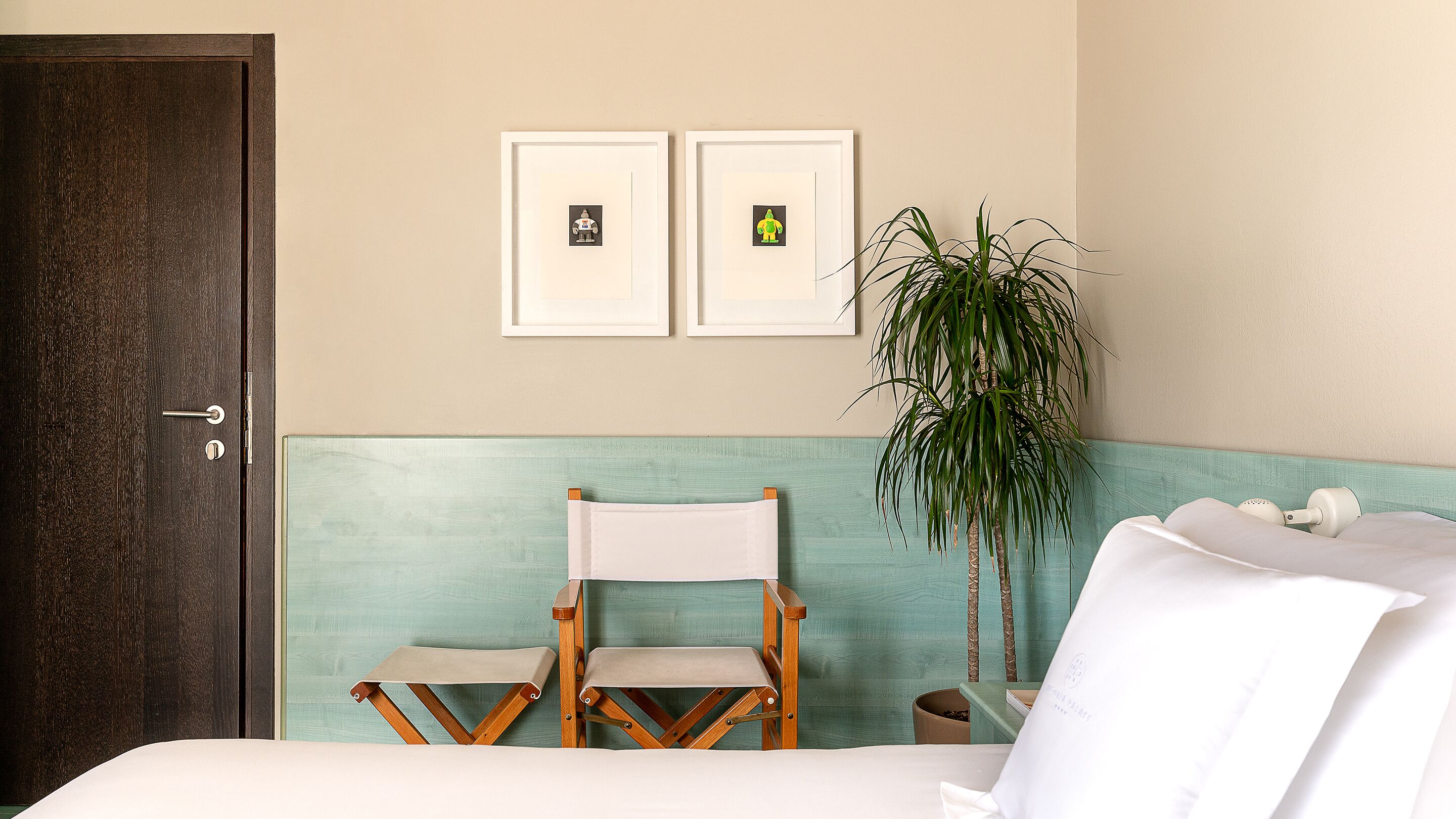 A hotel room with a bed, a wooden folding chair, a potted plant, and two framed pictures on the wall.