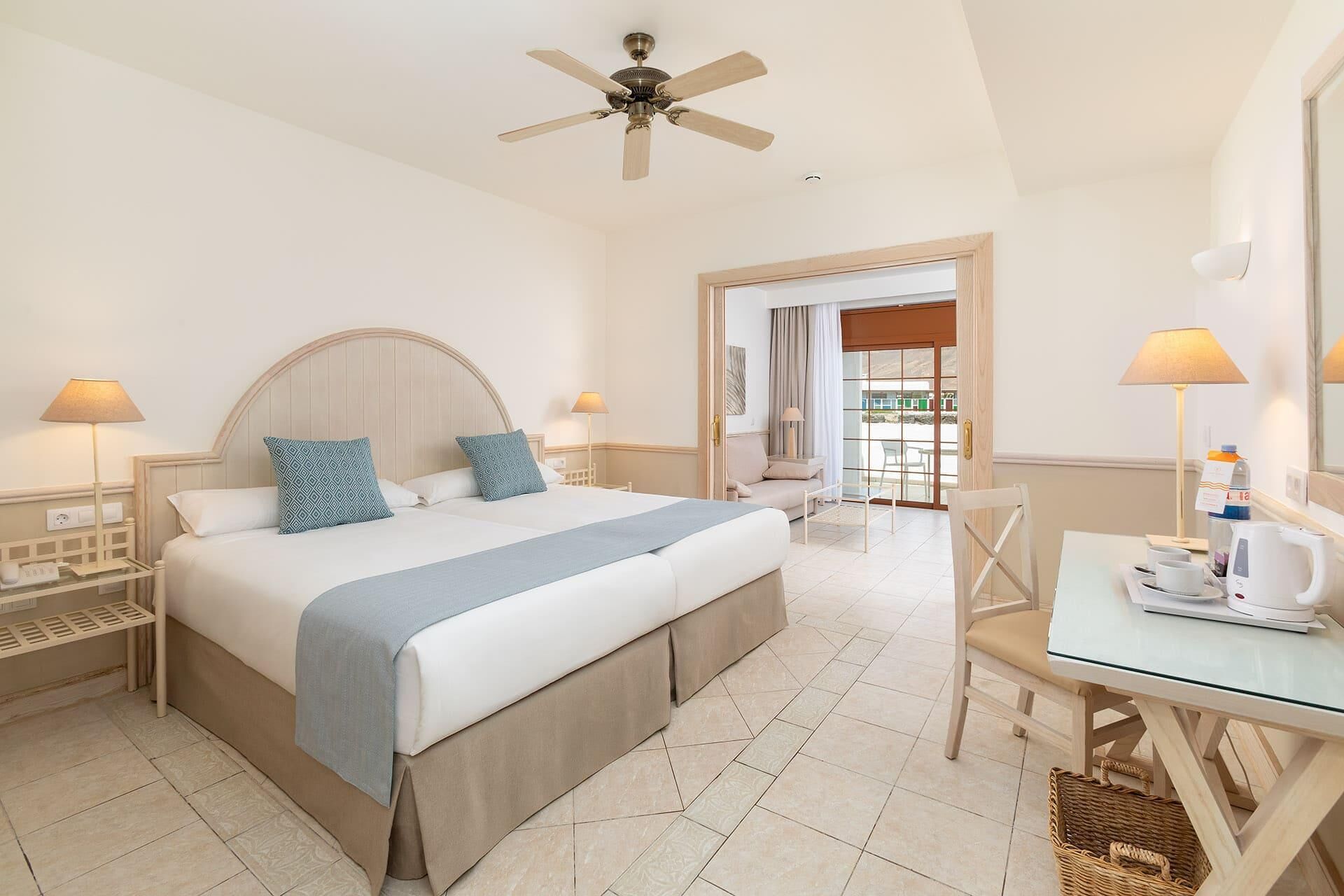 A hotel room with a bed, bedside tables, a desk, a chair, a ceiling fan, and a view through a glass door.
