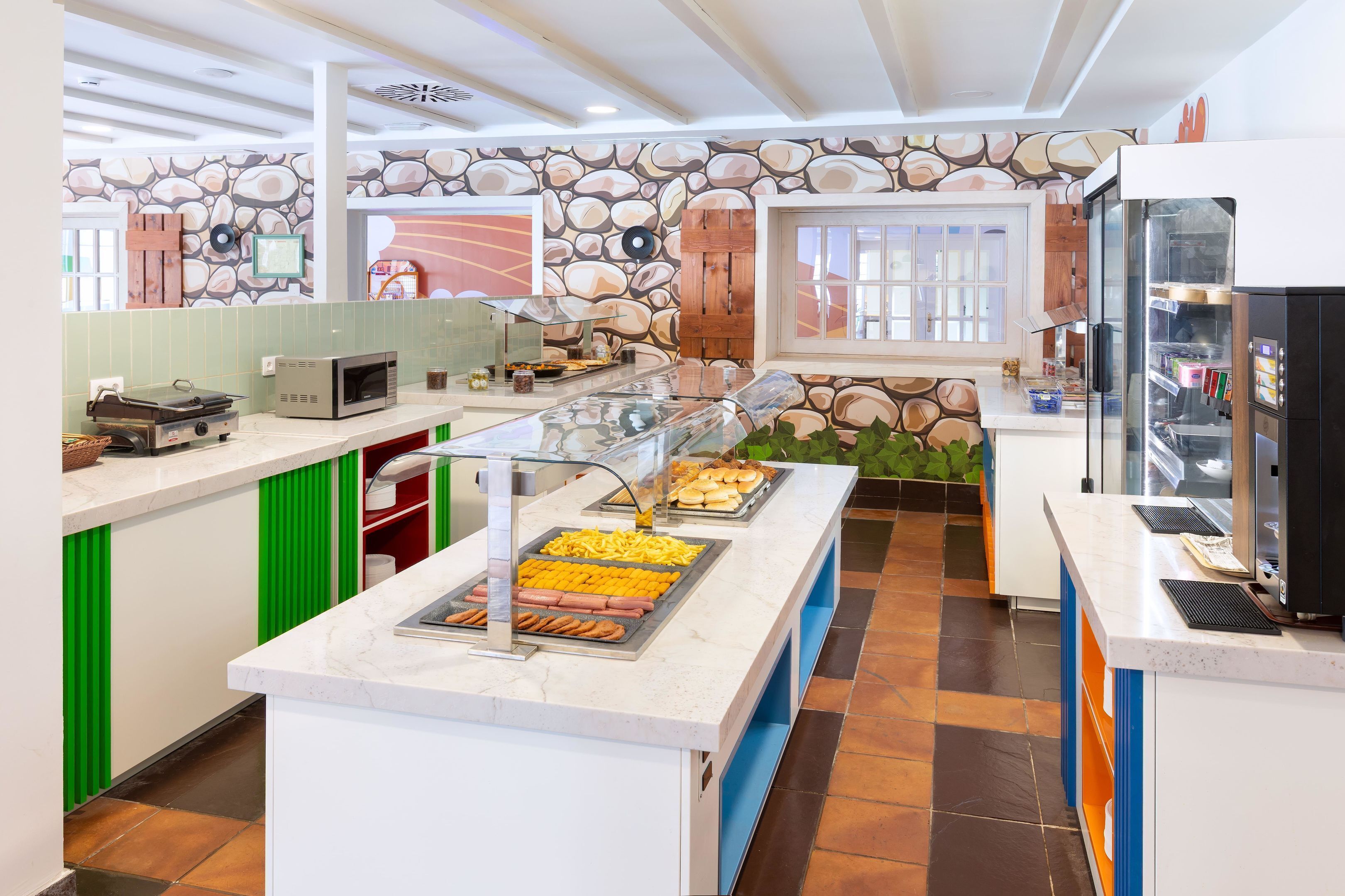 Espaço de café da manhã estilo buffet, com parede de pedra ao fundo, diversas opções de comida e cozinha moderna.