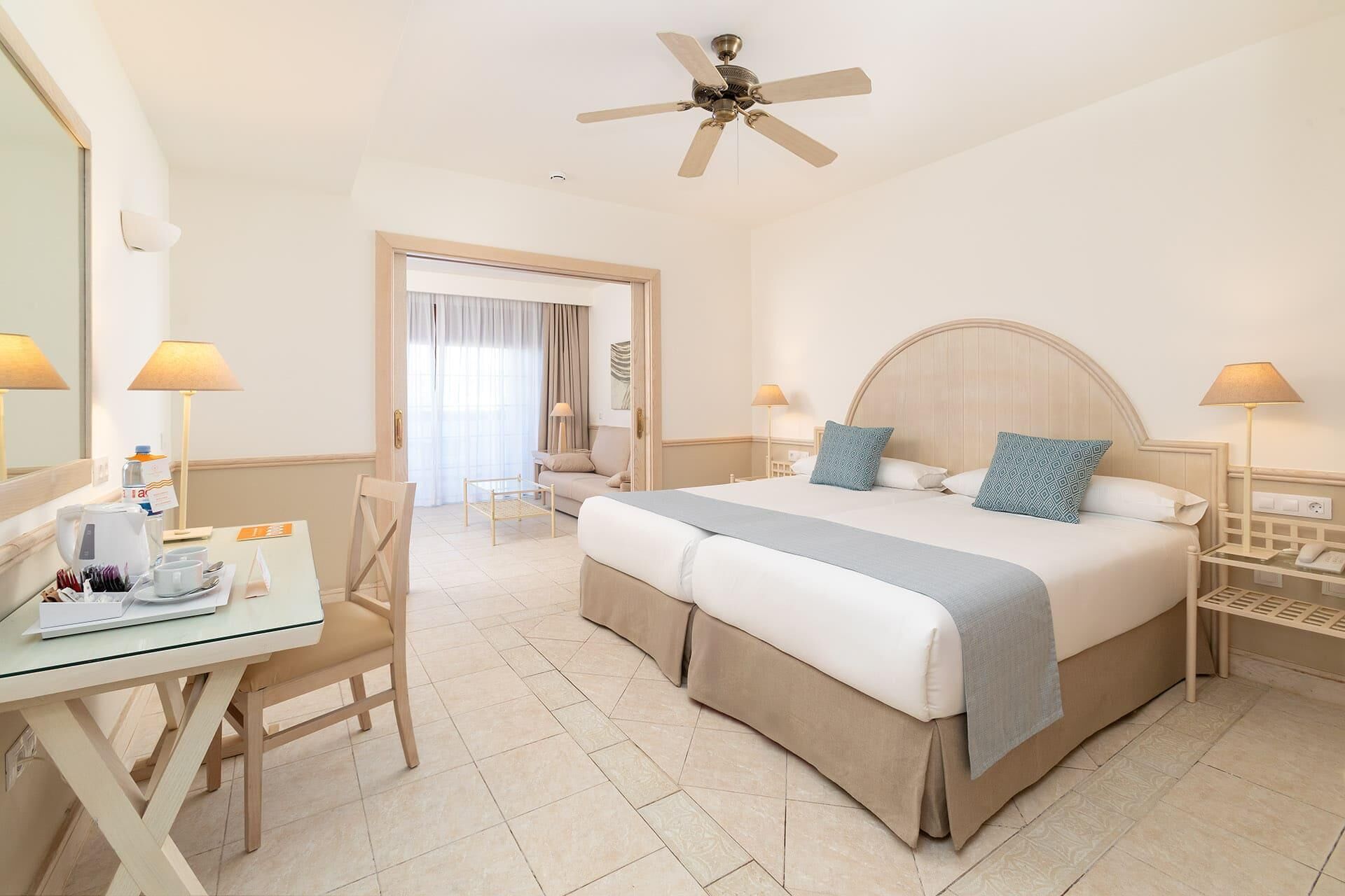 A hotel room with a bed, a desk, a chair, a ceiling fan, and a view of the outside.