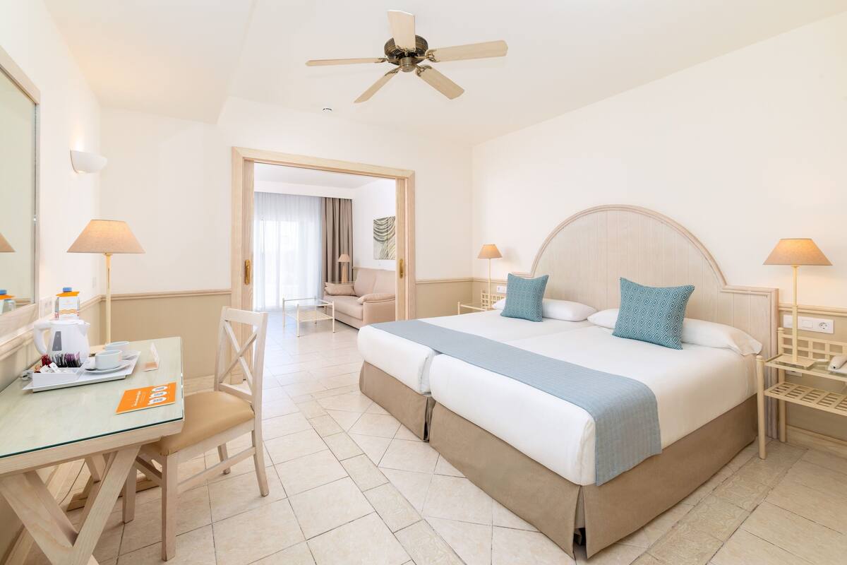 A hotel room with a bed, a desk with a chair, a table with a tray, a ceiling fan, and a door leading to another room.
