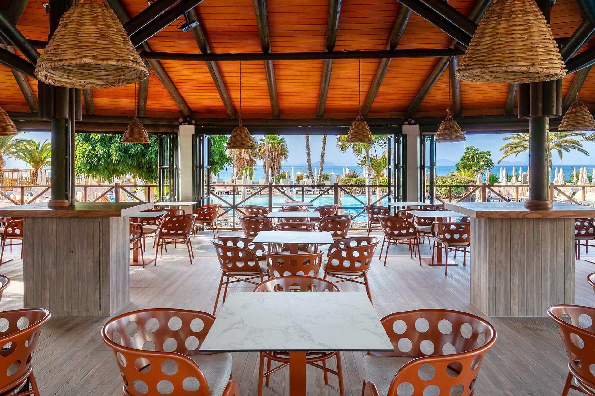 Un espacio de comedor al aire libre techado con mesas y sillas, con vistas a una piscina y al mar.