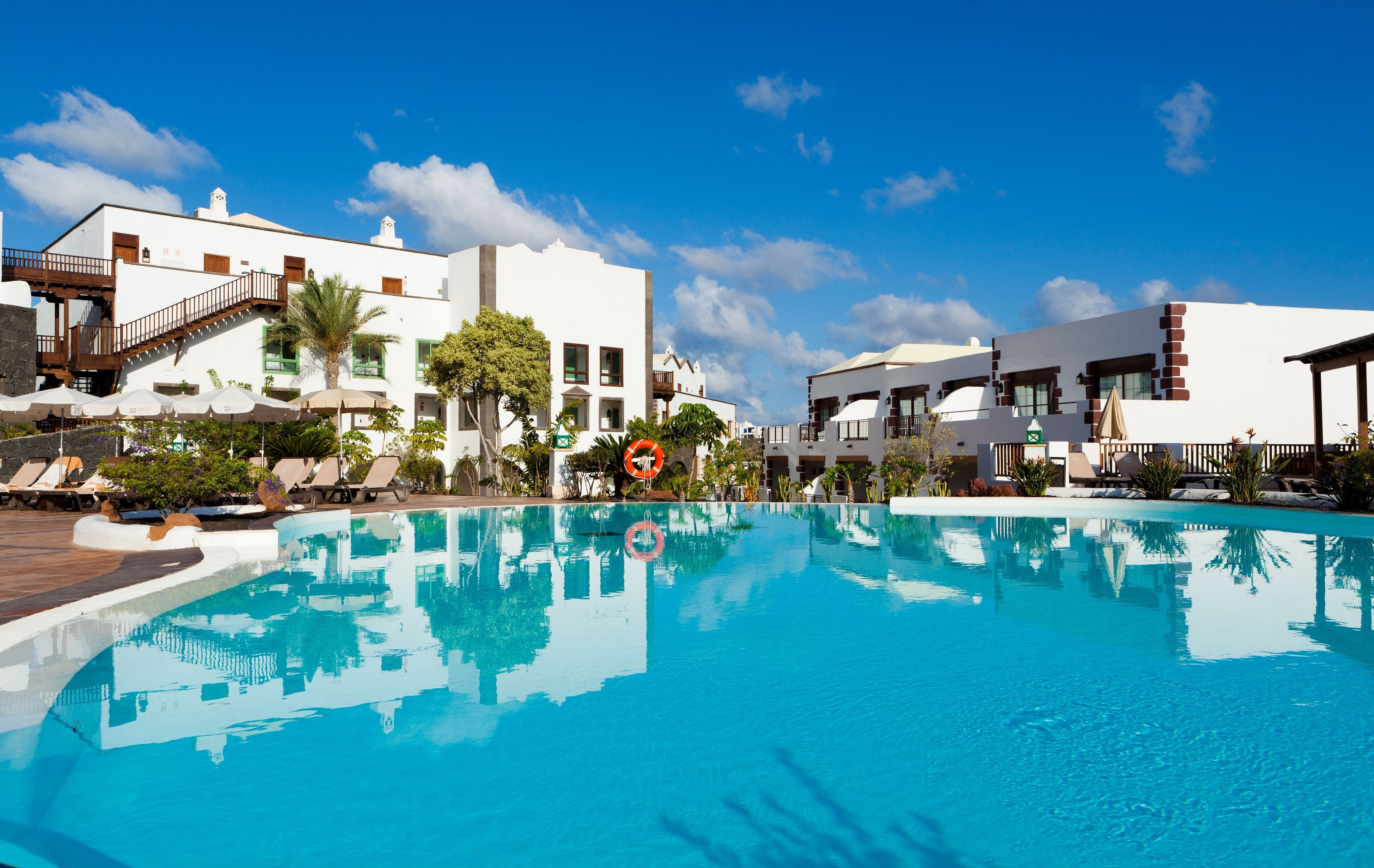 Une piscine entourée de chaises longues et de parasols, bordée de bâtiments blancs dotés de balcons, sous un ciel d’un bleu limpide.
