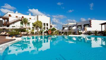 Una piscina con sillones y sombrillas, rodeada de edificios blancos con balcones y un cielo azul despejado.