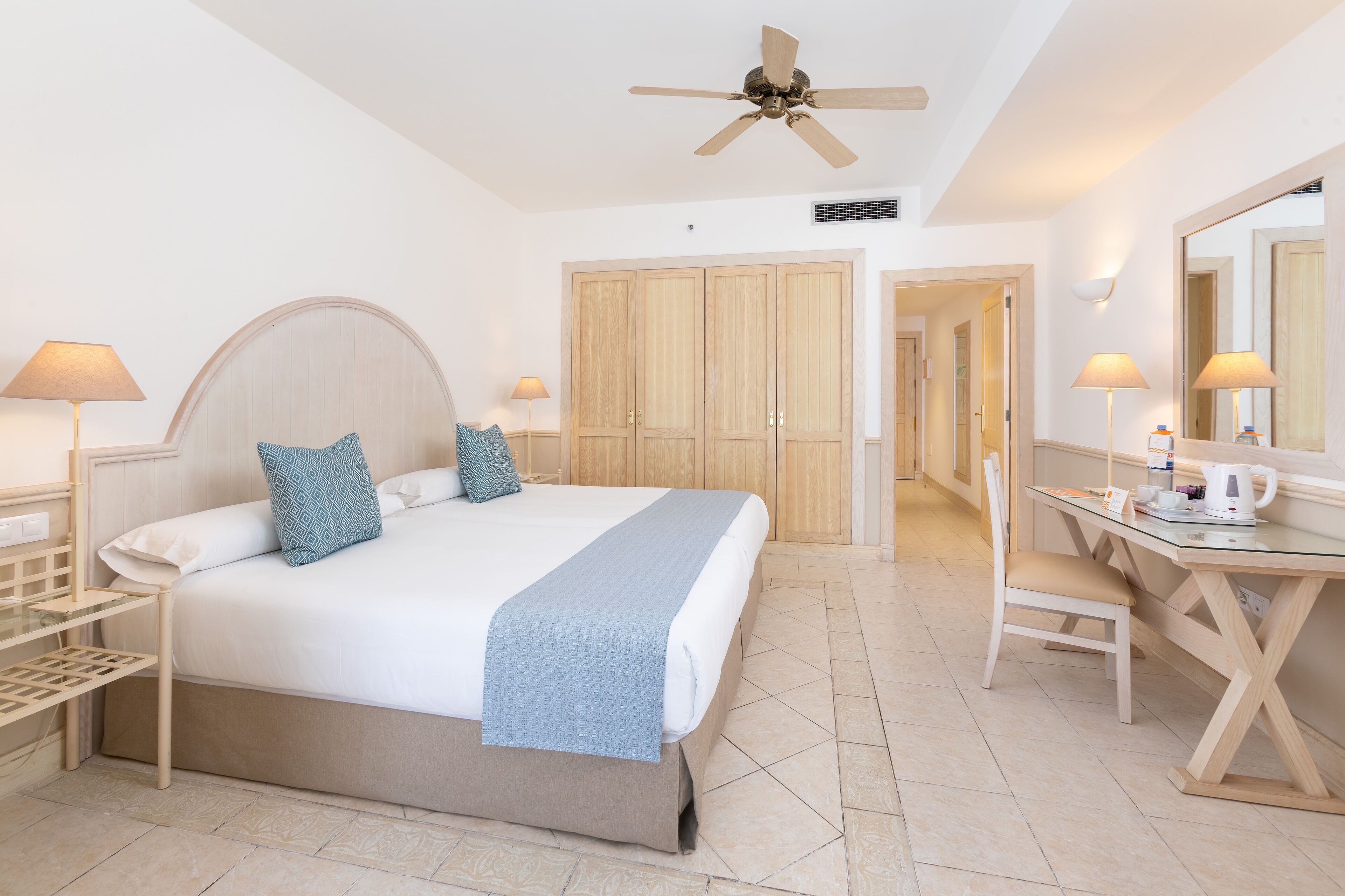 A hotel room with a bed, bedside tables, a desk, a chair, a ceiling fan, and a wardrobe.