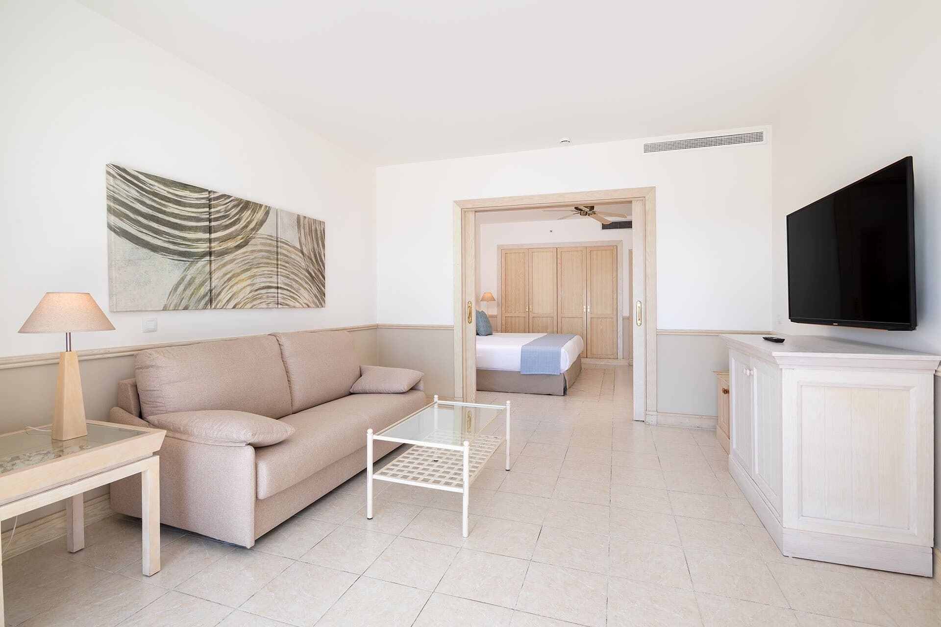 A modern hotel room with a beige sofa, a small white table, a TV, and a painting on the wall.