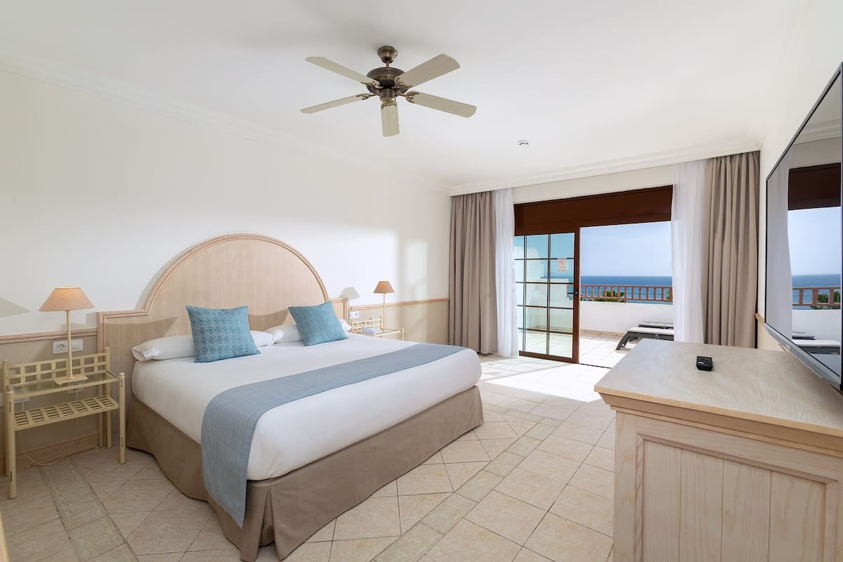 A hotel room with a bed, a desk, a ceiling fan, and a view of the ocean.