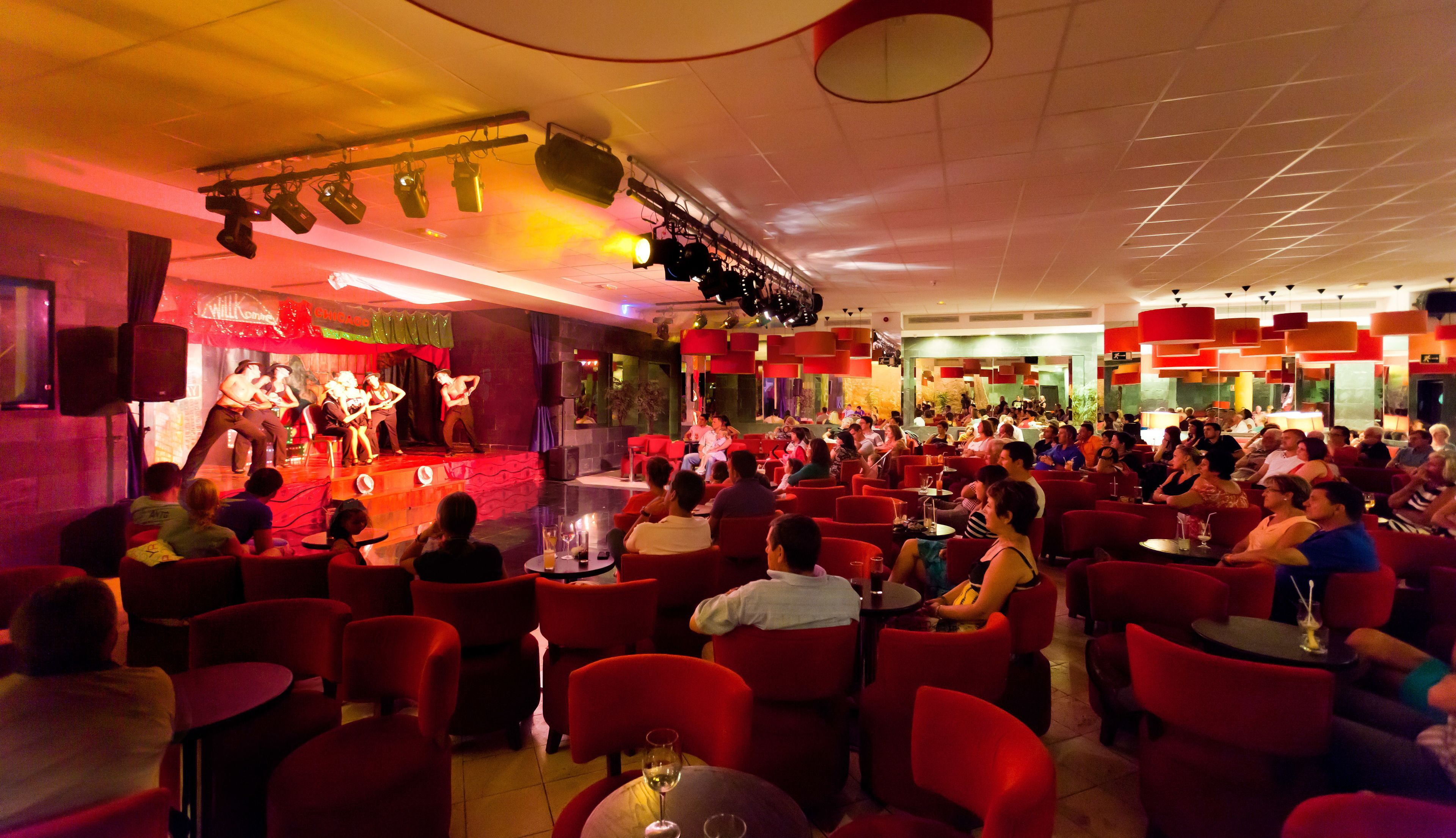 Un restaurante con sillas y mesas rojas, un escenario para espectáculos en vivo y una multitud de comensales.