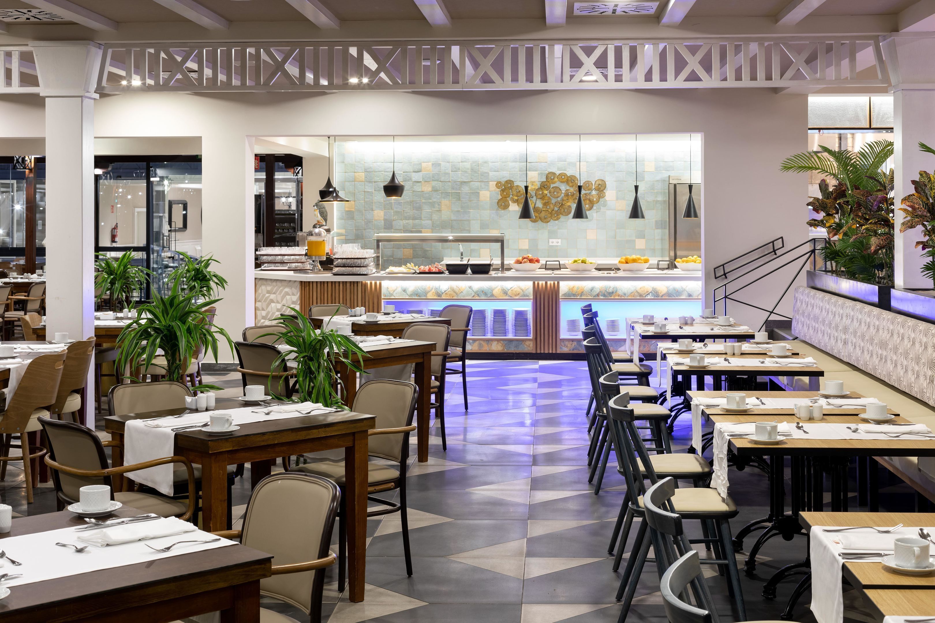 Un restaurant moderne doté d’un long comptoir, de nombreuses tables et des chaises. On y trouve des plantes en pot et un mur décoratif agrémenté de guirlandes lumineuses.