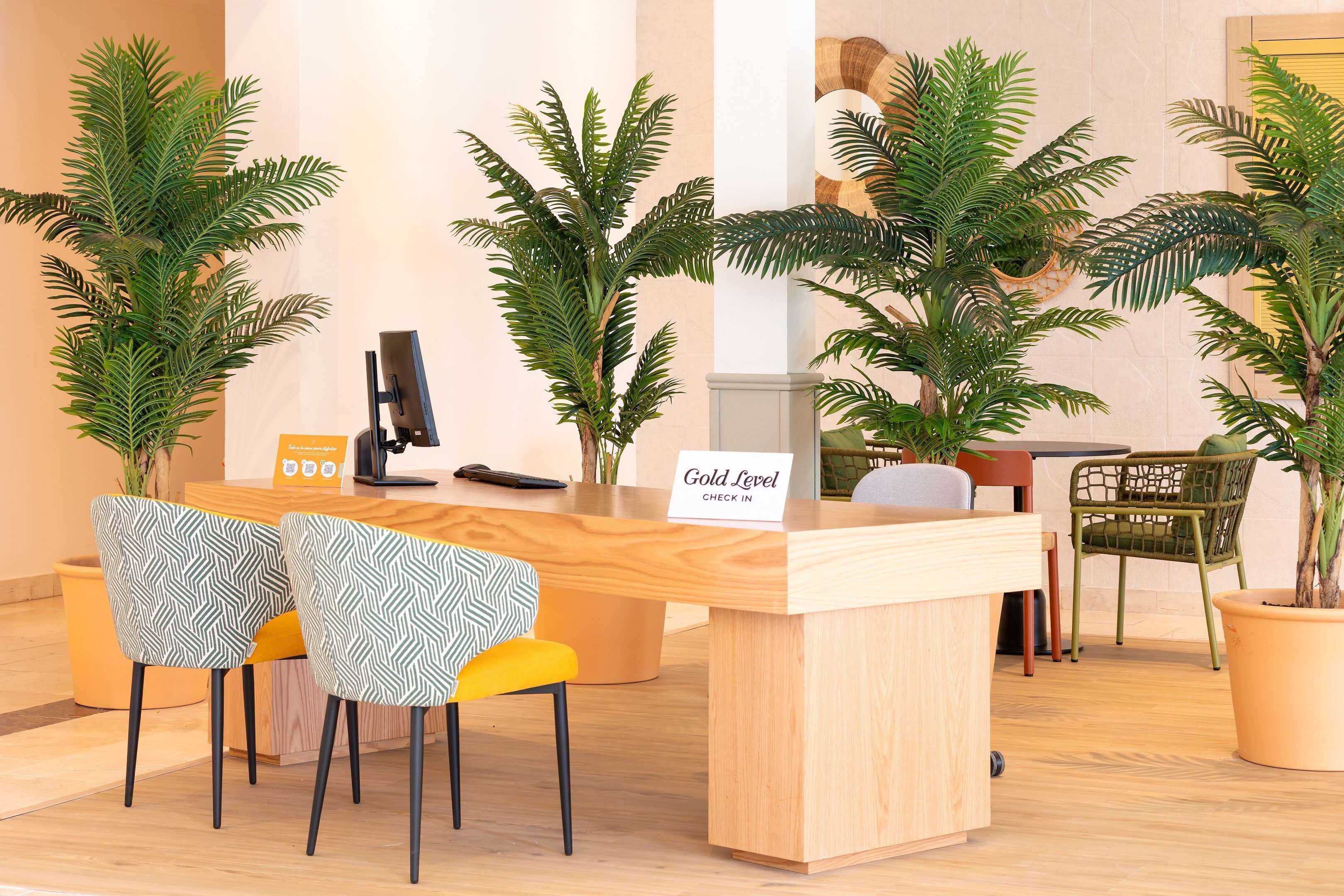 Mostrador de recepción con un cartel que dice 'Gold Level Check In', rodeado de plantas en macetas y dos sillas estampadas.