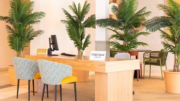 Mostrador de recepción con un cartel que dice 'Gold Level Check In', rodeado de plantas en macetas y dos sillas estampadas.
