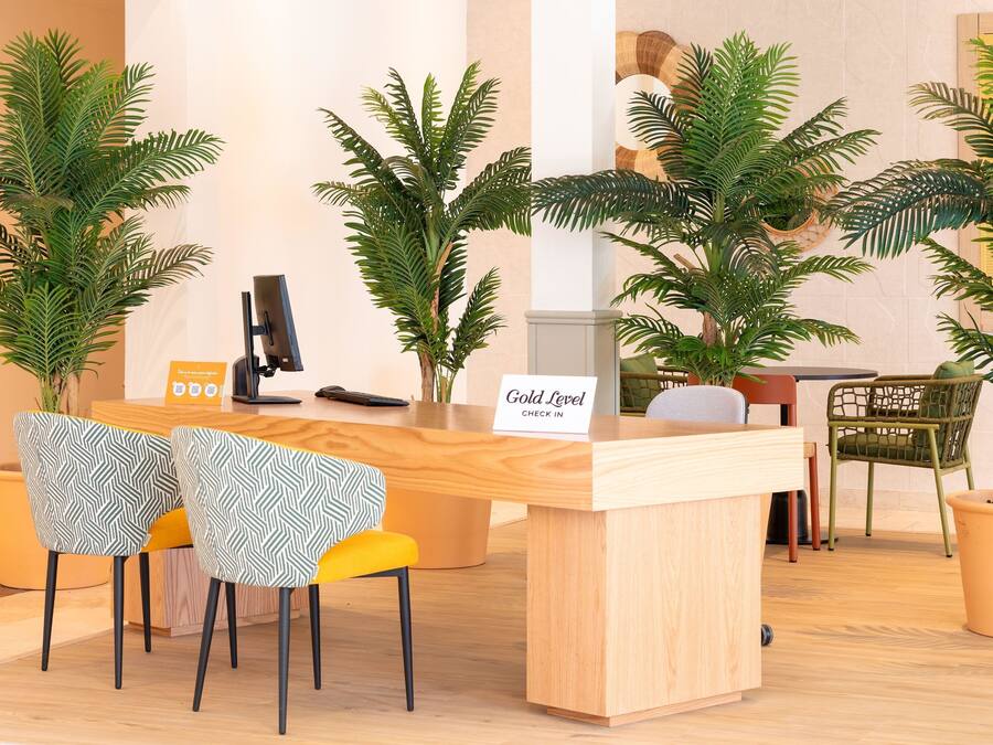Mostrador de recepción con un cartel que dice 'Gold Level Check In', rodeado de plantas en macetas y dos sillas estampadas.