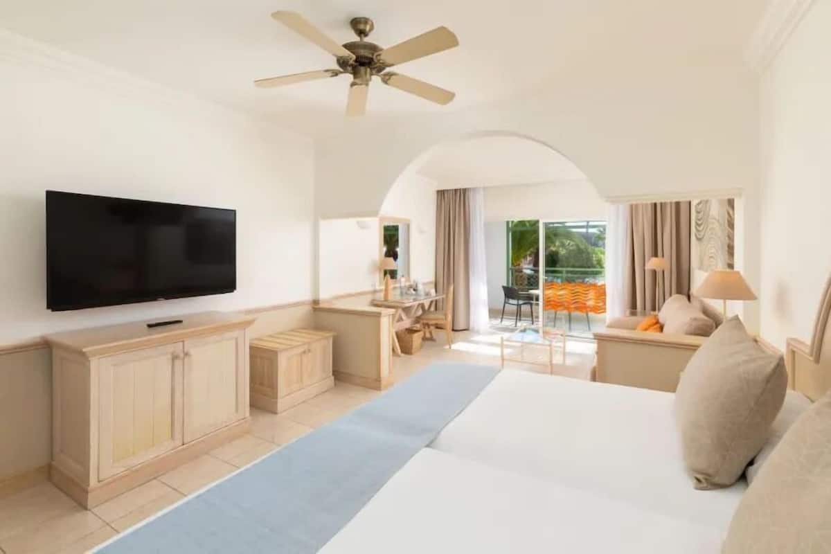 A hotel room with a large bed, a flat-screen TV, a ceiling fan, a desk, and a balcony with outdoor seating.