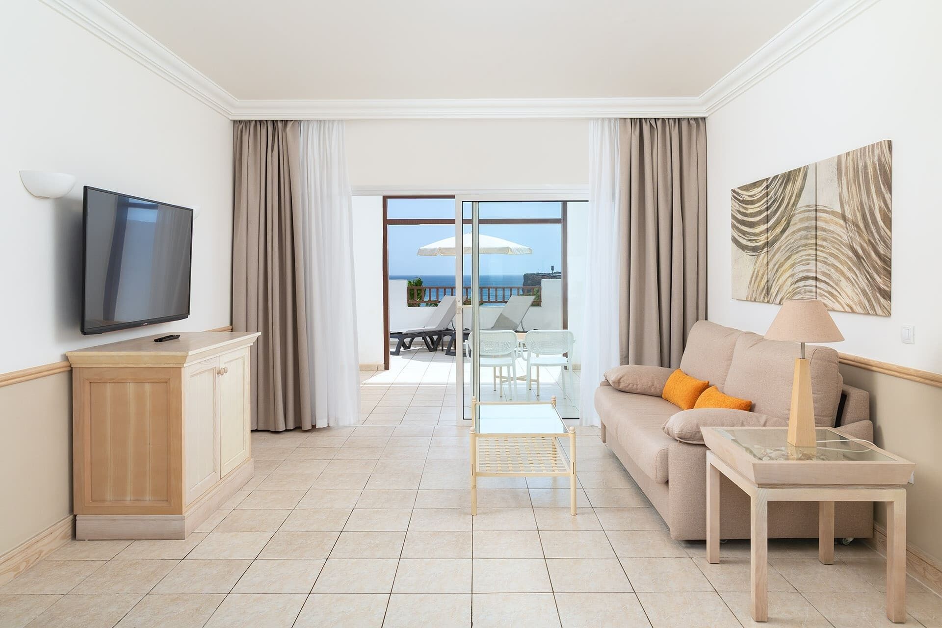 Quarto de hotel moderno com televisão de tela plana, sofá com almofadas, mesa pequena e divisória de vidro que dá acesso a uma área externa com assentos e vista para o mar.