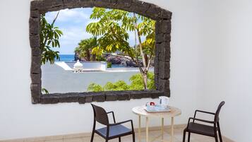 Una habitación con ventana enmarcada en piedra que da a una playa con palmeras y un edificio blanco. Hay dos sillas y una pequeña mesa redonda con una taza y platillo blancos.