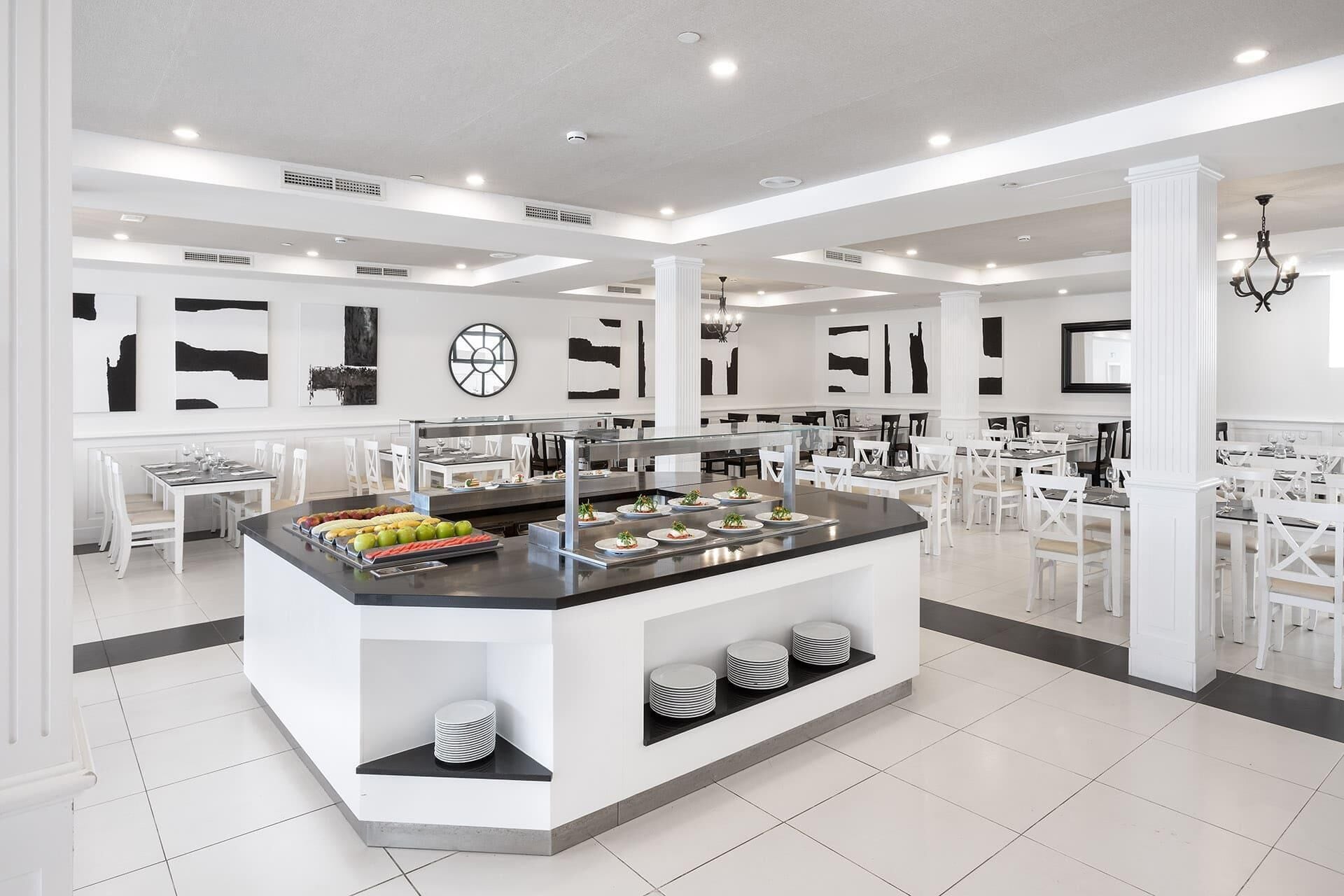 Un comedor moderno y espacioso con una isla central que ofrece un sistema tipo bufé, rodeado de mesas y sillas blancas.