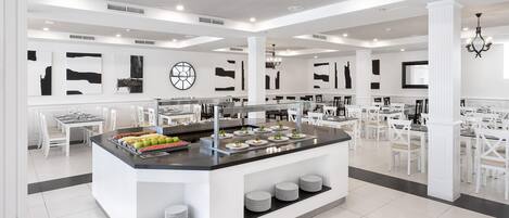 Un comedor moderno y espacioso con una isla central que ofrece un sistema tipo bufé, rodeado de mesas y sillas blancas.