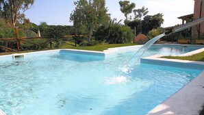 Una piscina rectangular con agua azul cristalina, un tobogán acuático y una zona verde circundante con árboles.