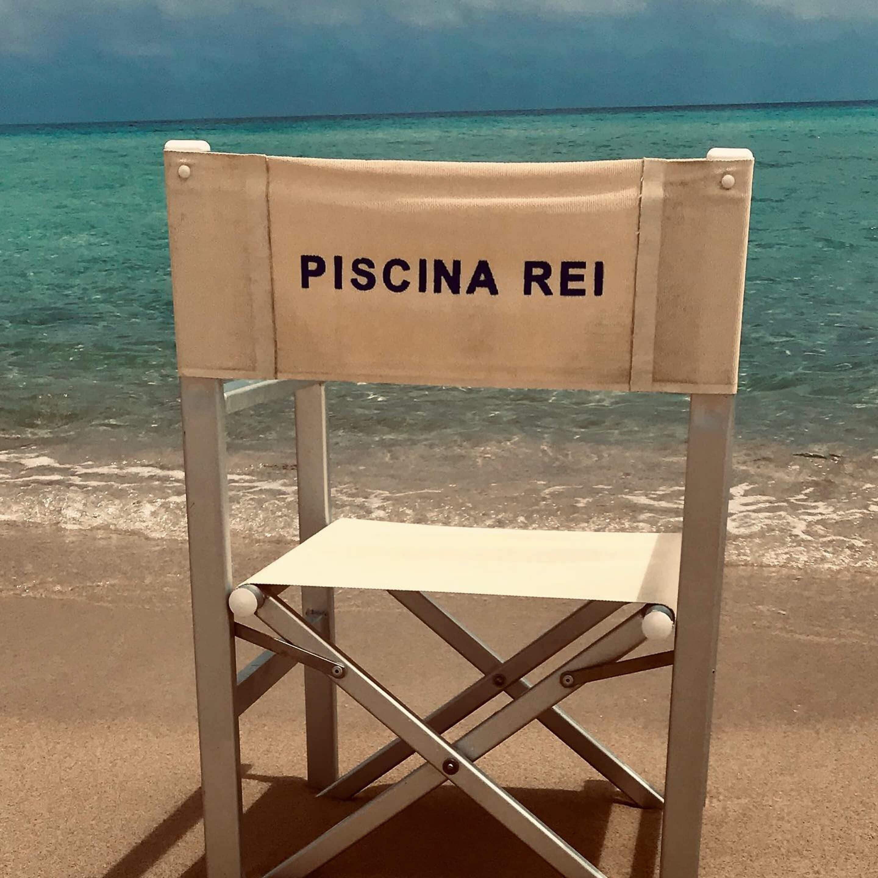 Una silla de playa con el texto "PISCINA REI" en el respaldo, ubicada en una playa de arena con agua turquesa cristalina al fondo.