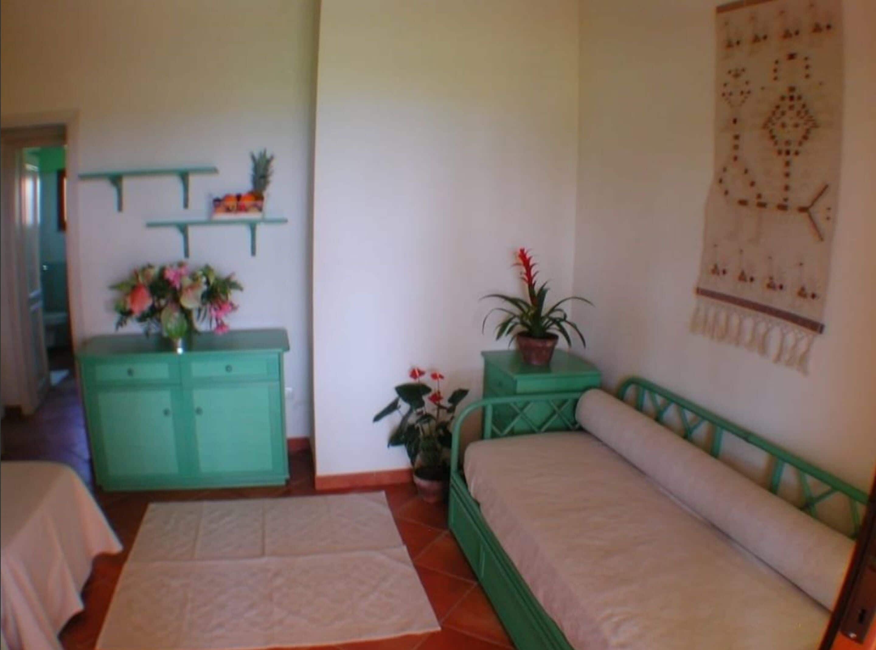Una habitación con una cama, una mesita con una planta, un aparador con un espejo y un adorno en la pared.