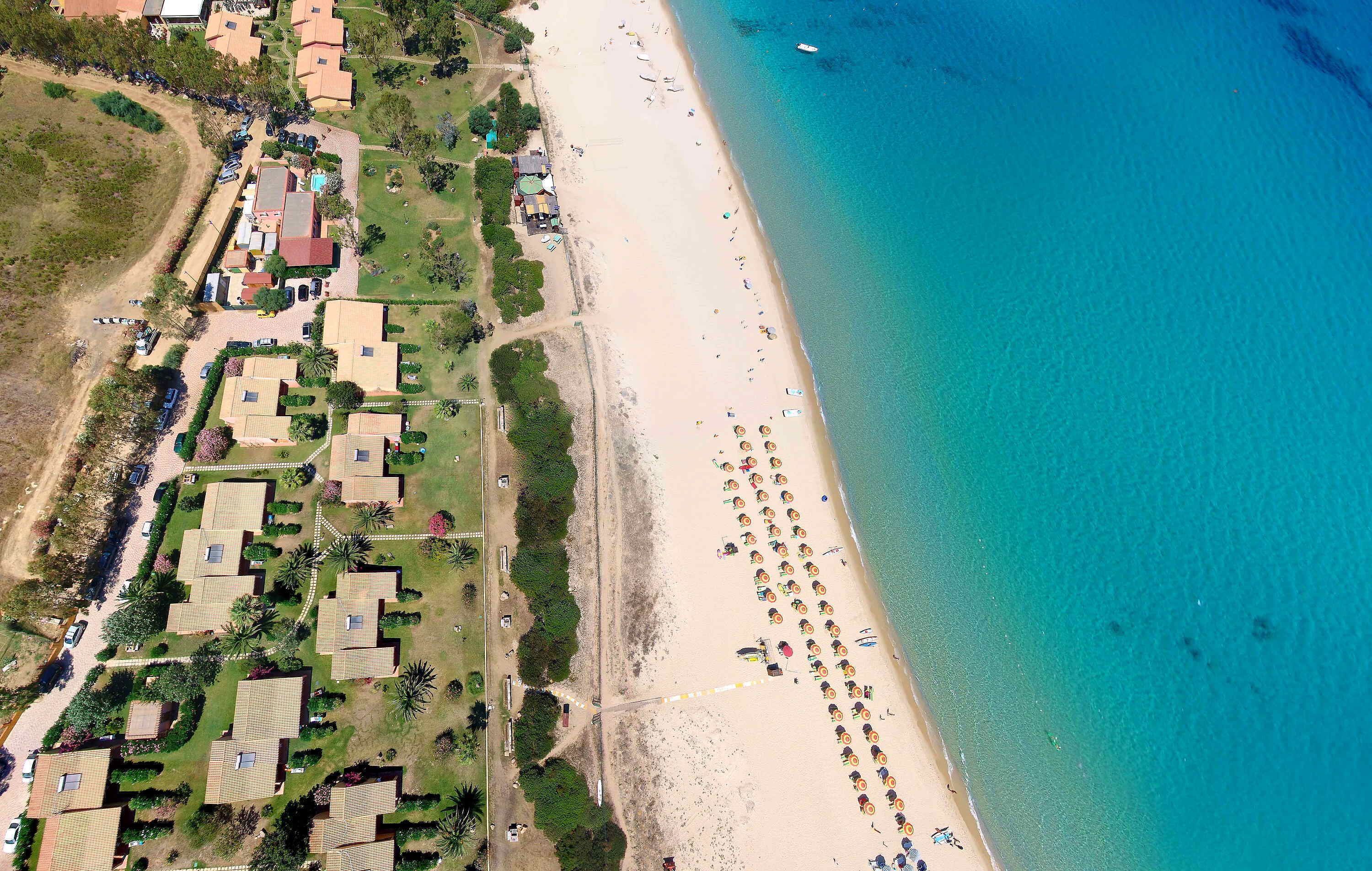 An aerial view of a coastal area with a beach, a row of houses, and a parking lot.