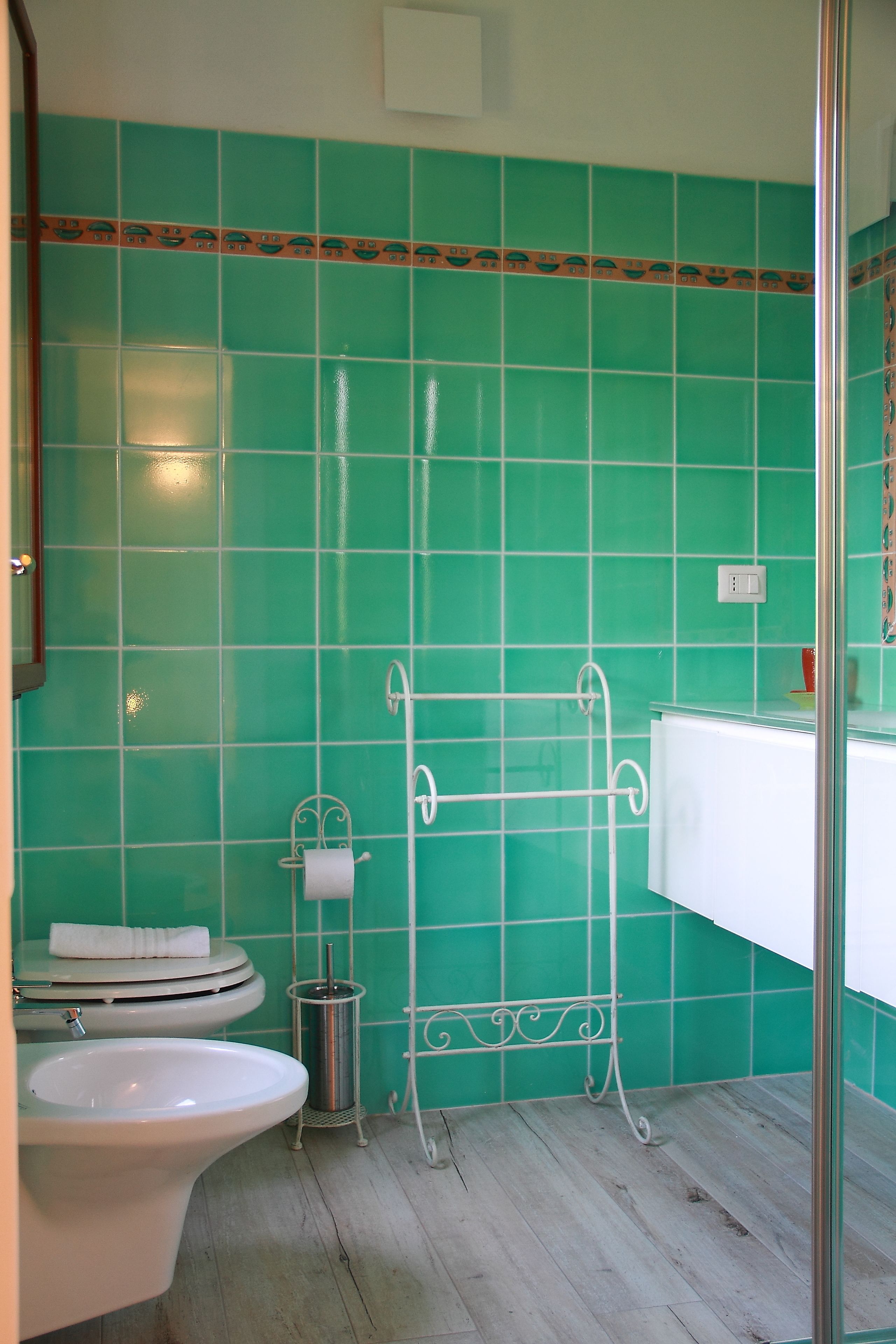 Une salle de bain avec des murs recouverts de carreaux verts, des toilettes blanches, un lavabo blanc et un porte-serviettes en métal.