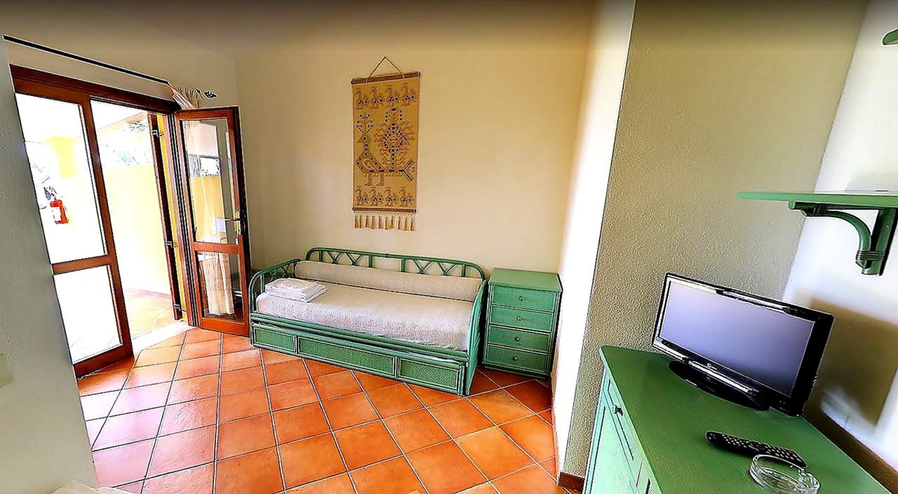 A small room with a green bed, a green dresser, a television on a stand, and a wall hanging.