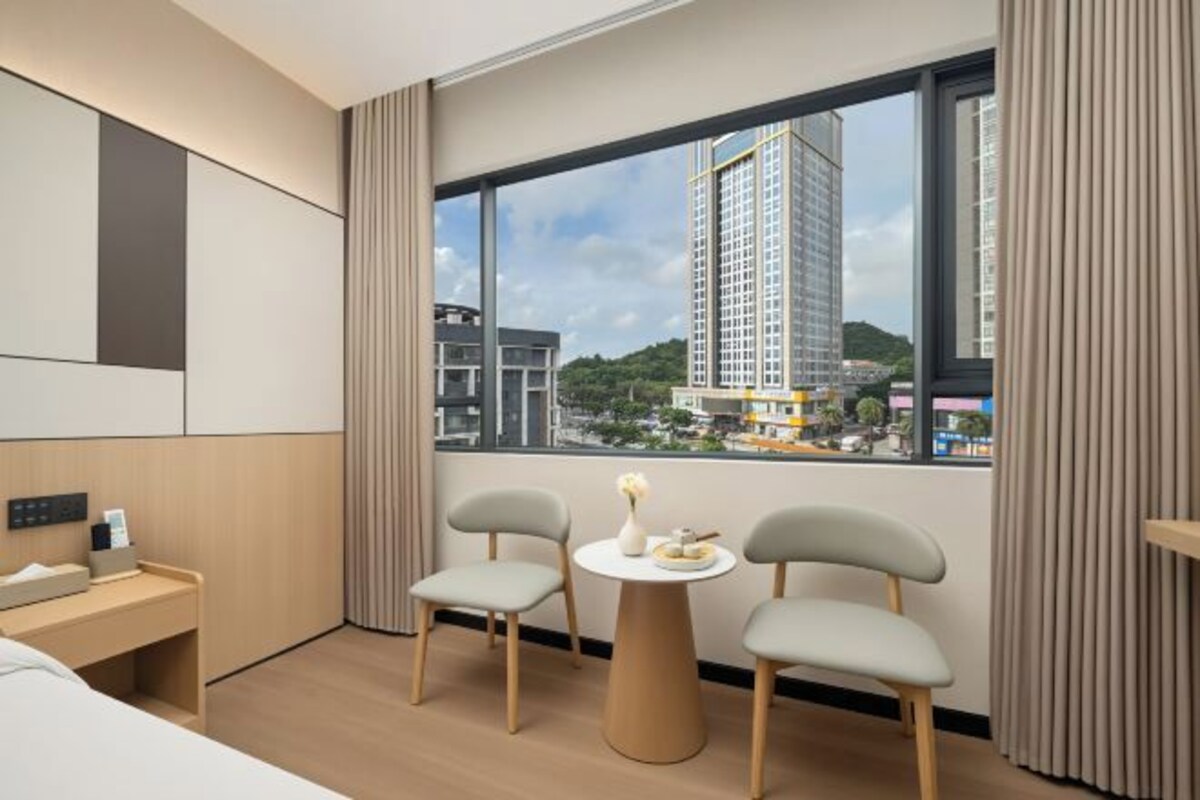 Une chambre d’hôtel avec un lit, un bureau, deux chaises, une petite table et une grande fenêtre donnant sur des immeubles et une colline.