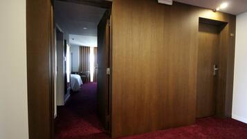 A hotel hallway with wooden doors, a red carpet, and a glimpse of a room with a bed and curtains in the background.