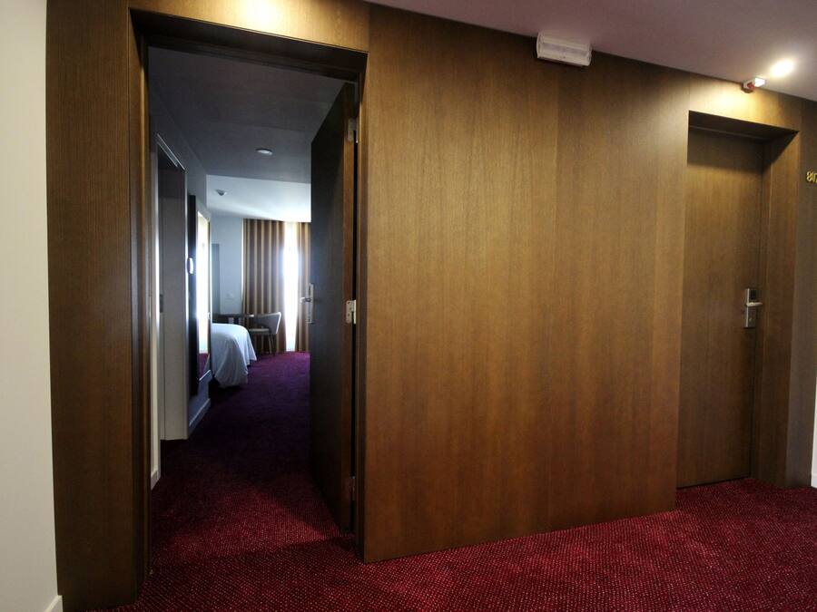 A hotel hallway with wooden doors, a red carpet, and a glimpse of a room with a bed and curtains in the background.