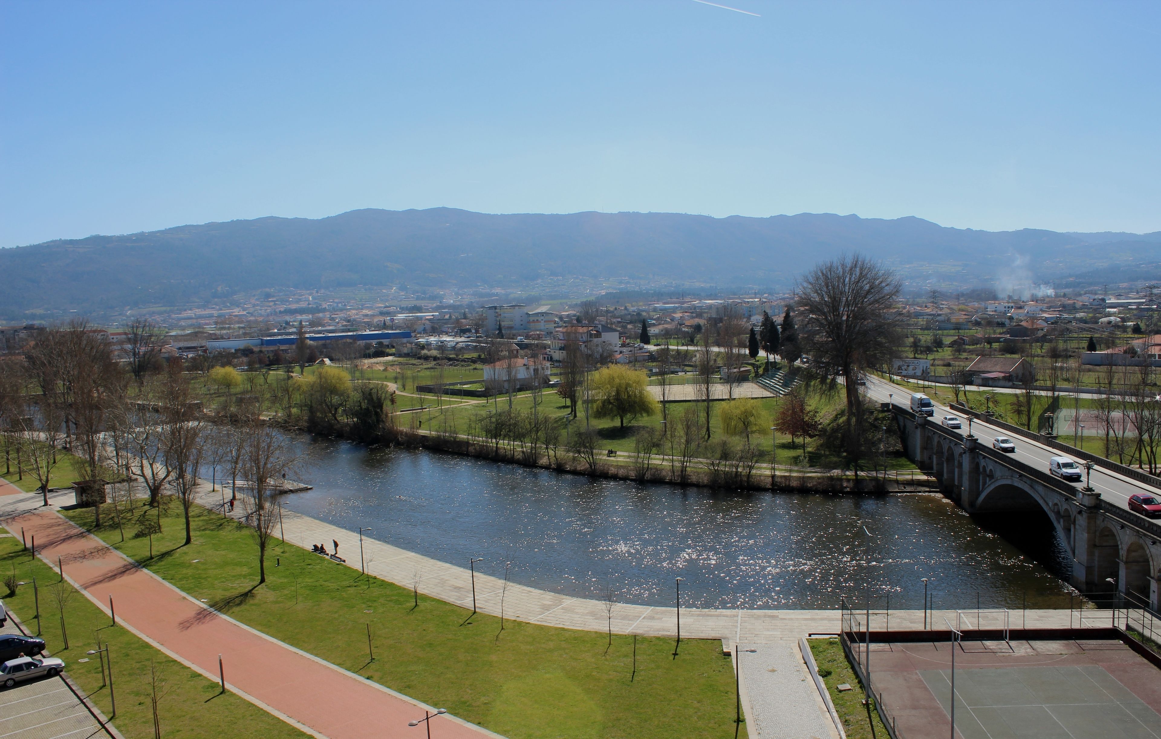A river with a bridge, a park with a walking path, and a distant cityscape.