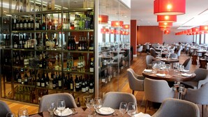 A restaurant with a wine cellar, multiple dining tables, and red hanging lights.