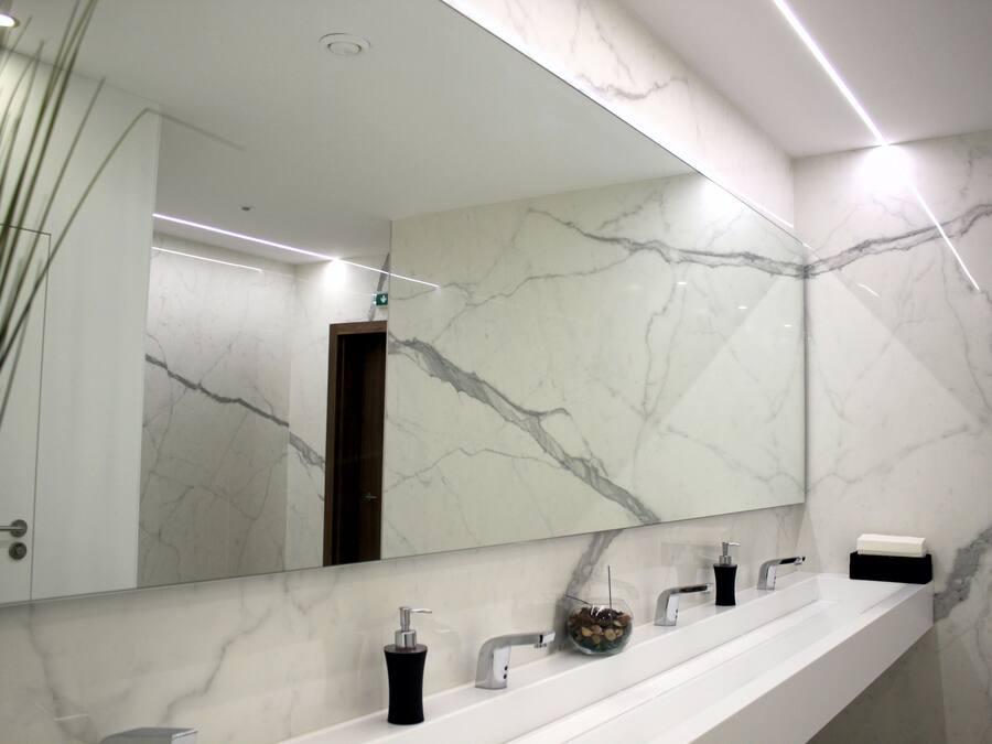 A modern bathroom with a marble wall, a large mirror, and a long white countertop with multiple sinks and a soap dispenser.